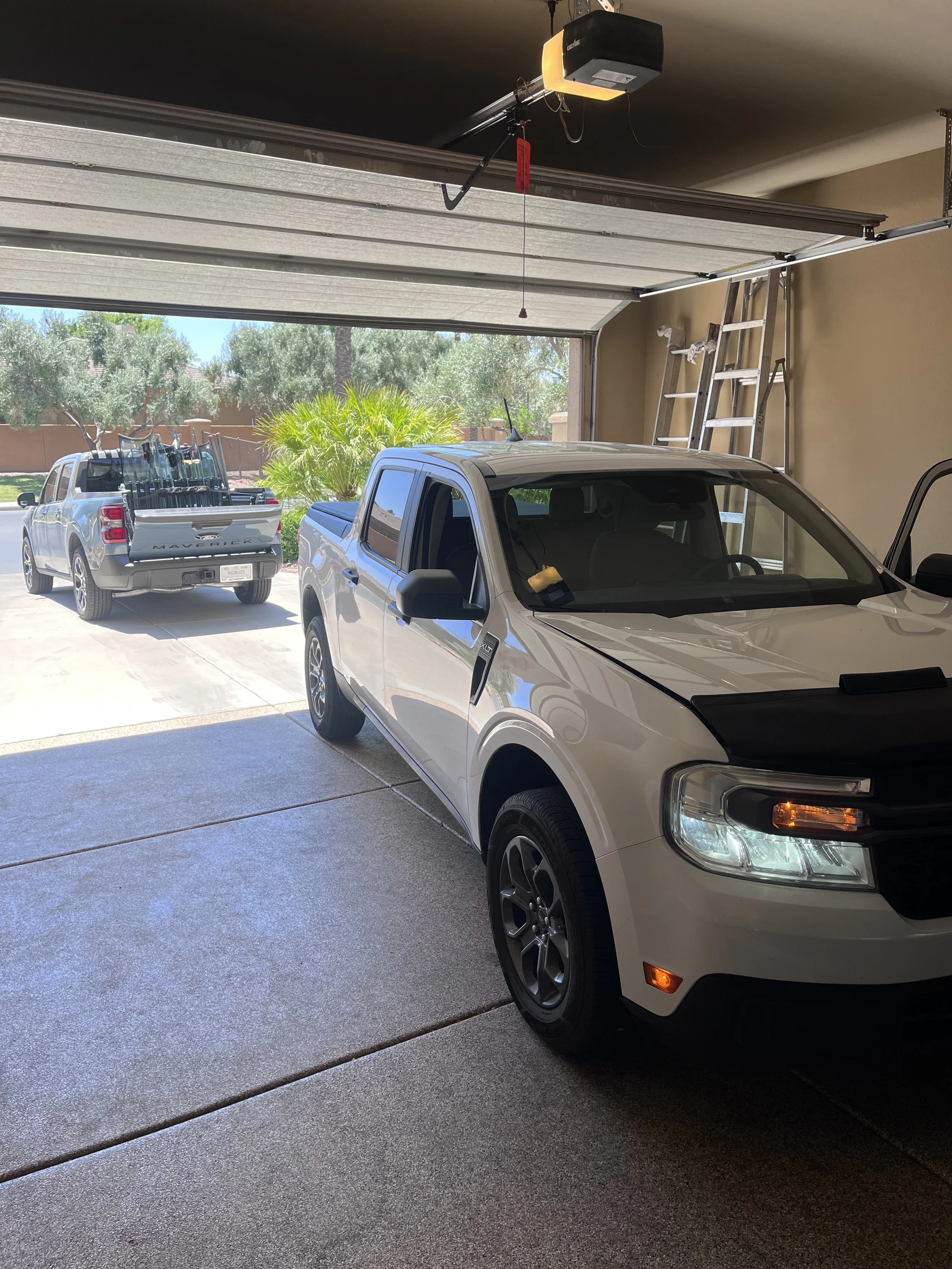 Inside view of a two-car garage with the garage door open, showing two pickup trucks parked outside, with a ladder leaning against the right wall and a garage door opener mounted on the ceiling.