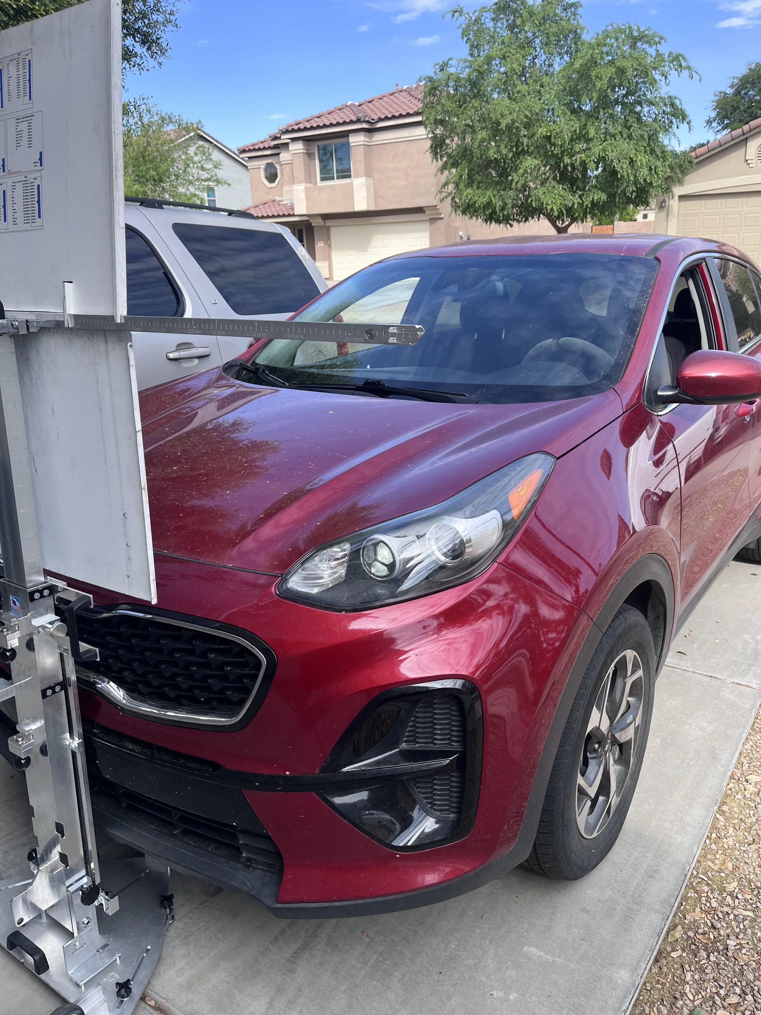 A red compact SUV parked in a residential driveway, with a metallic measuring device extending from a nearby metal structure in front of it, and a house and trees in the background.