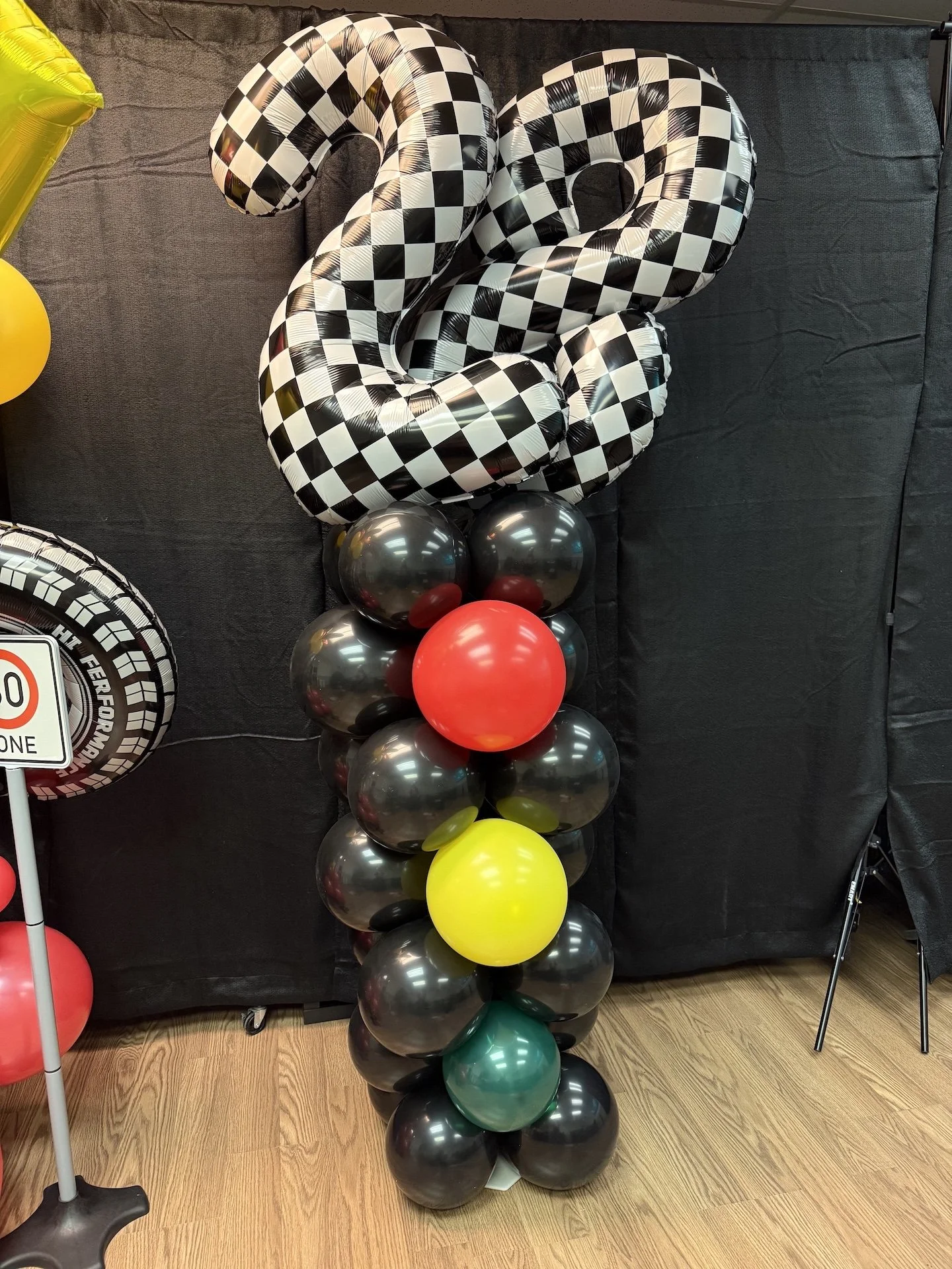 A decorative balloon arrangement featuring a large checkered trophy number '2' at the top, surrounded by assorted black, red, yellow, and green balloons, against a dark background and wooden floor.