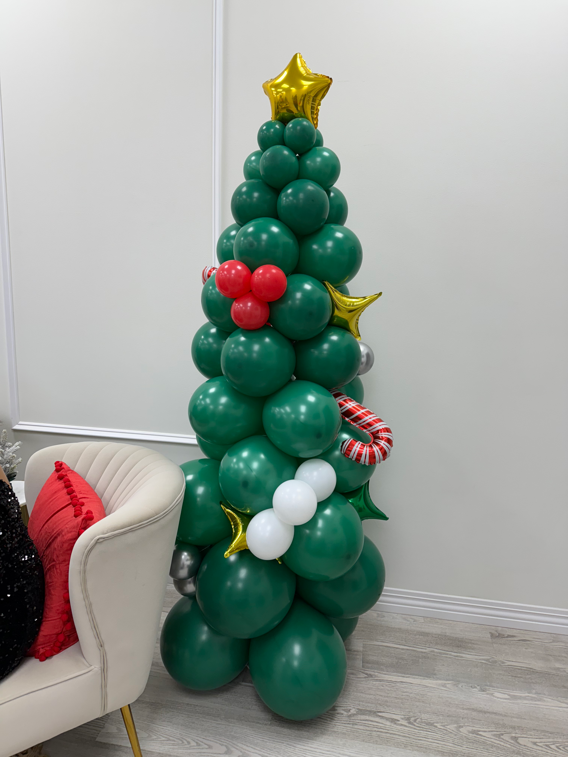 Decorative Christmas tree made of green balloons with gold star balloons on top, red and white striped balloon candy cane, white balloons, and small red balloon ornaments, positioned next to a white chair with a red cushion.