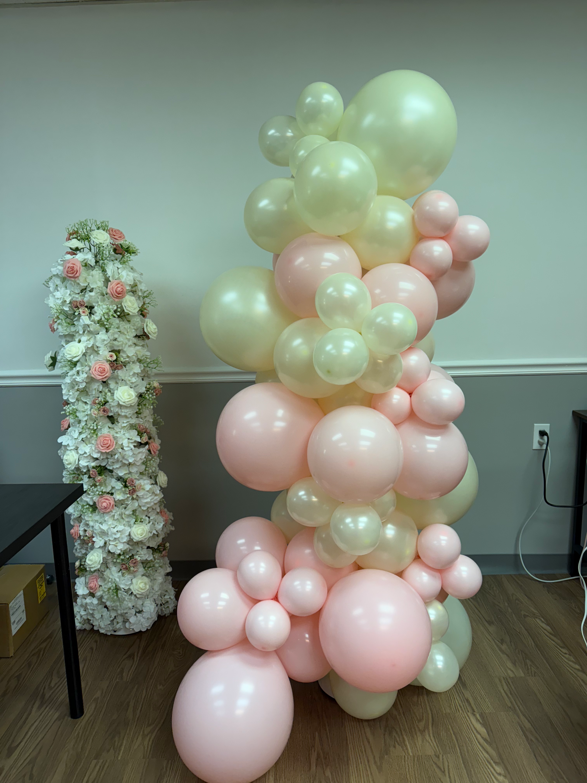 A decorative arrangement of pastel-colored balloons in pink, cream, and light green, with a floral display of white and pink roses and flowers in the background.