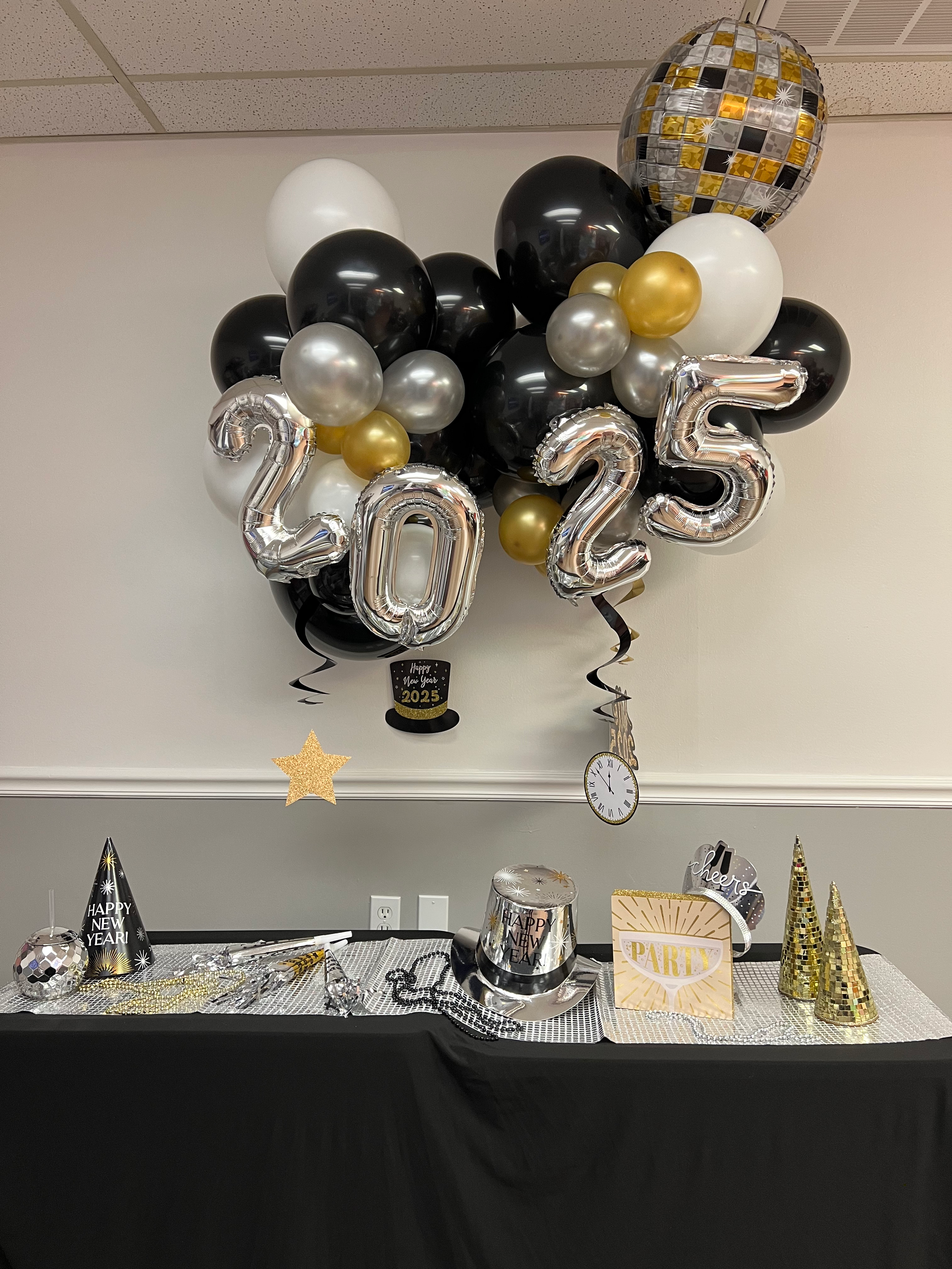 Decorative balloons with silver, black, white, and gold colors, shaped as the numbers 2025, are floating against a ceiling. The table below features New Year’s Eve party decorations including a “Happy New Year 2025” black top hat, a “Happy New Year”
