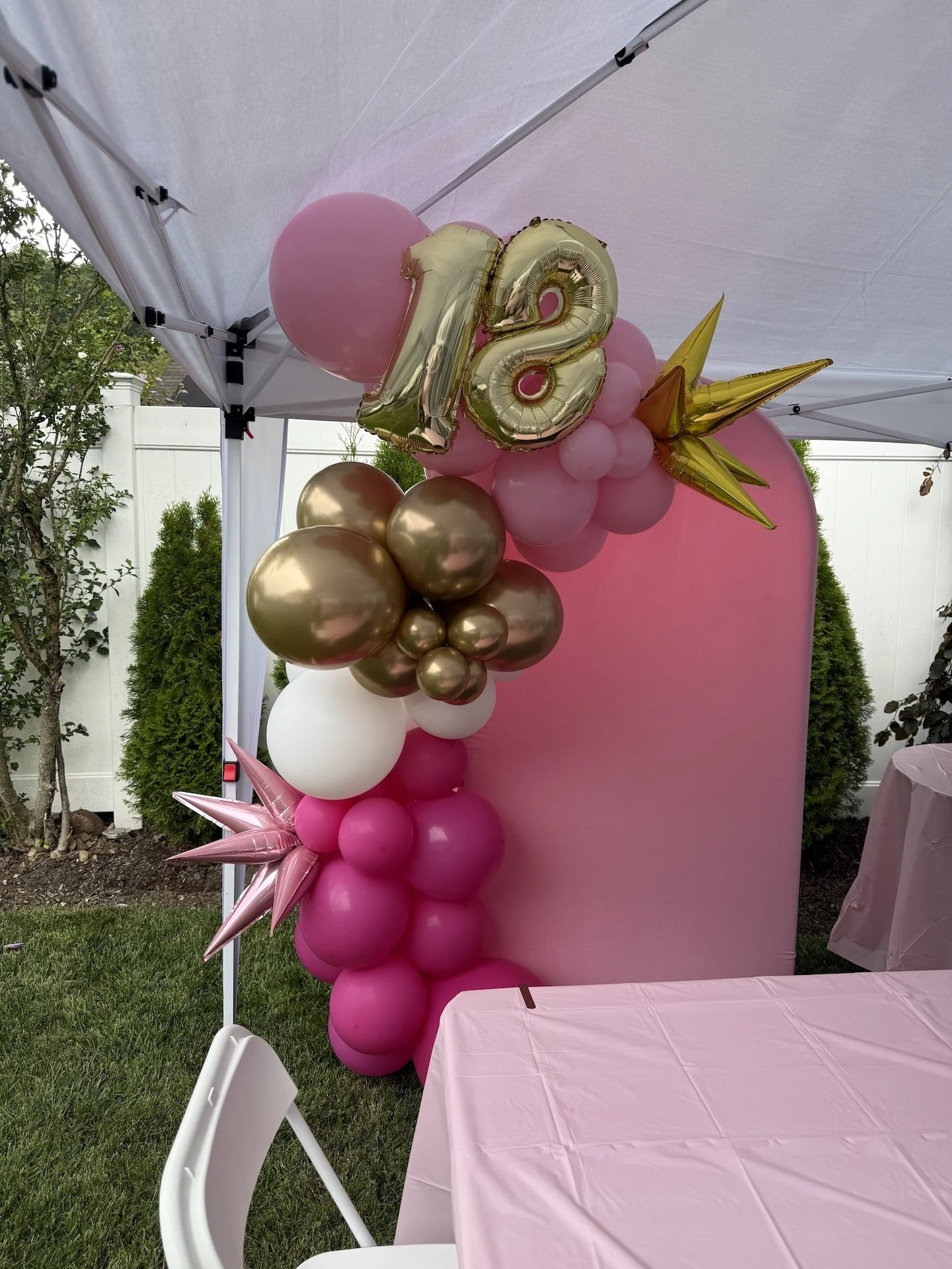 Balloon decoration with gold and pink balloons, including a gold '18' balloon, gold star-shaped balloons, pink, white, and gold round balloons, set up for a celebration outdoors on grass.