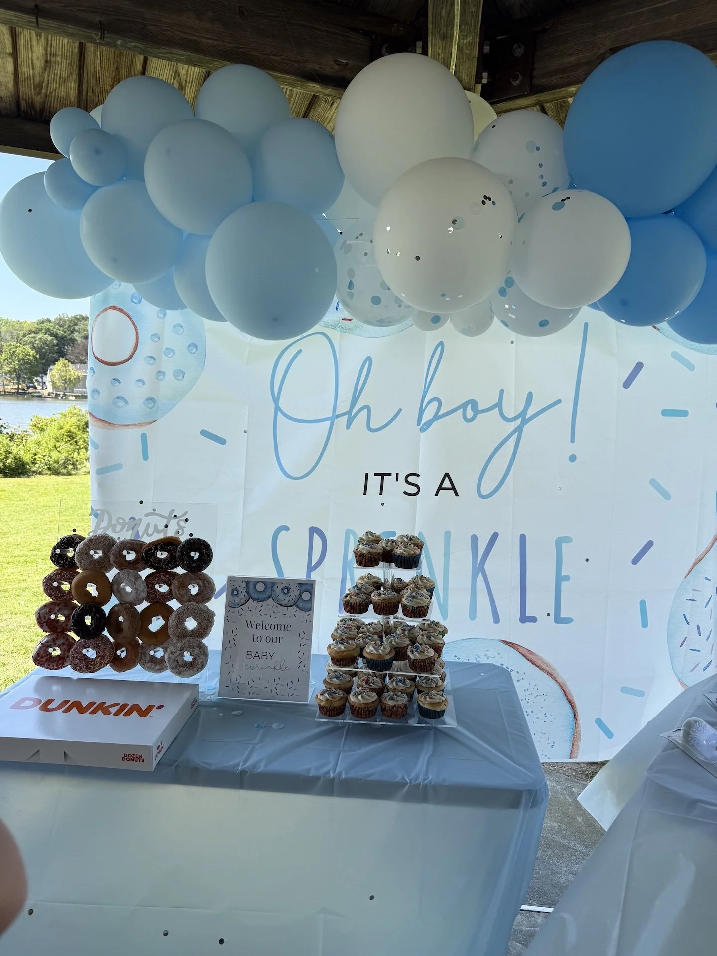 Decorative table with cupcakes and donut treats under white and blue balloons at a baby shower with a backdrop saying, 'Oh boy! It's a sprinkle.'