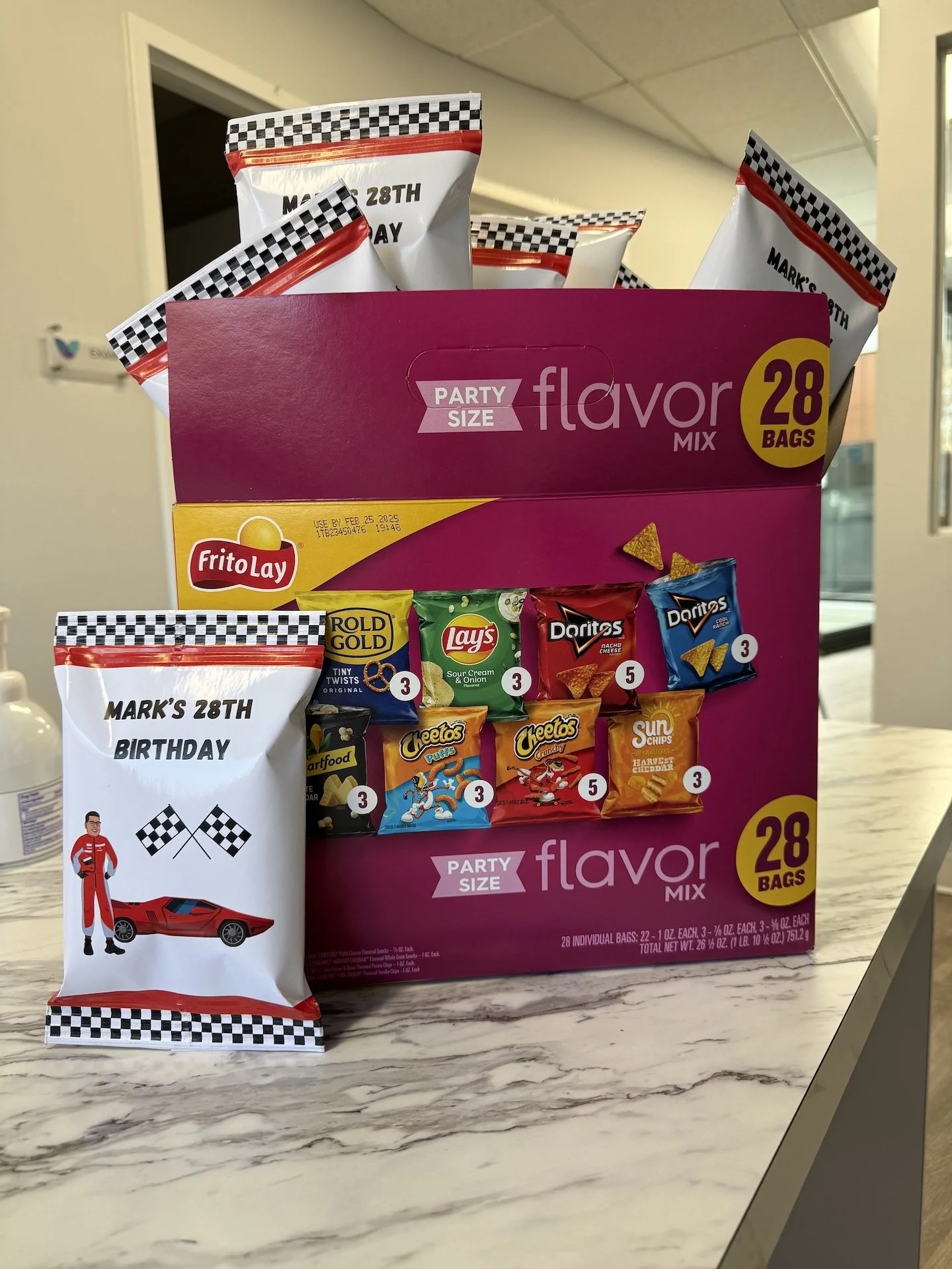 A party snack box containing 28 bags of chips and snack-sized treats, with a single birthday-themed snack bag placed in front, featuring a racing car and checkered flag design.