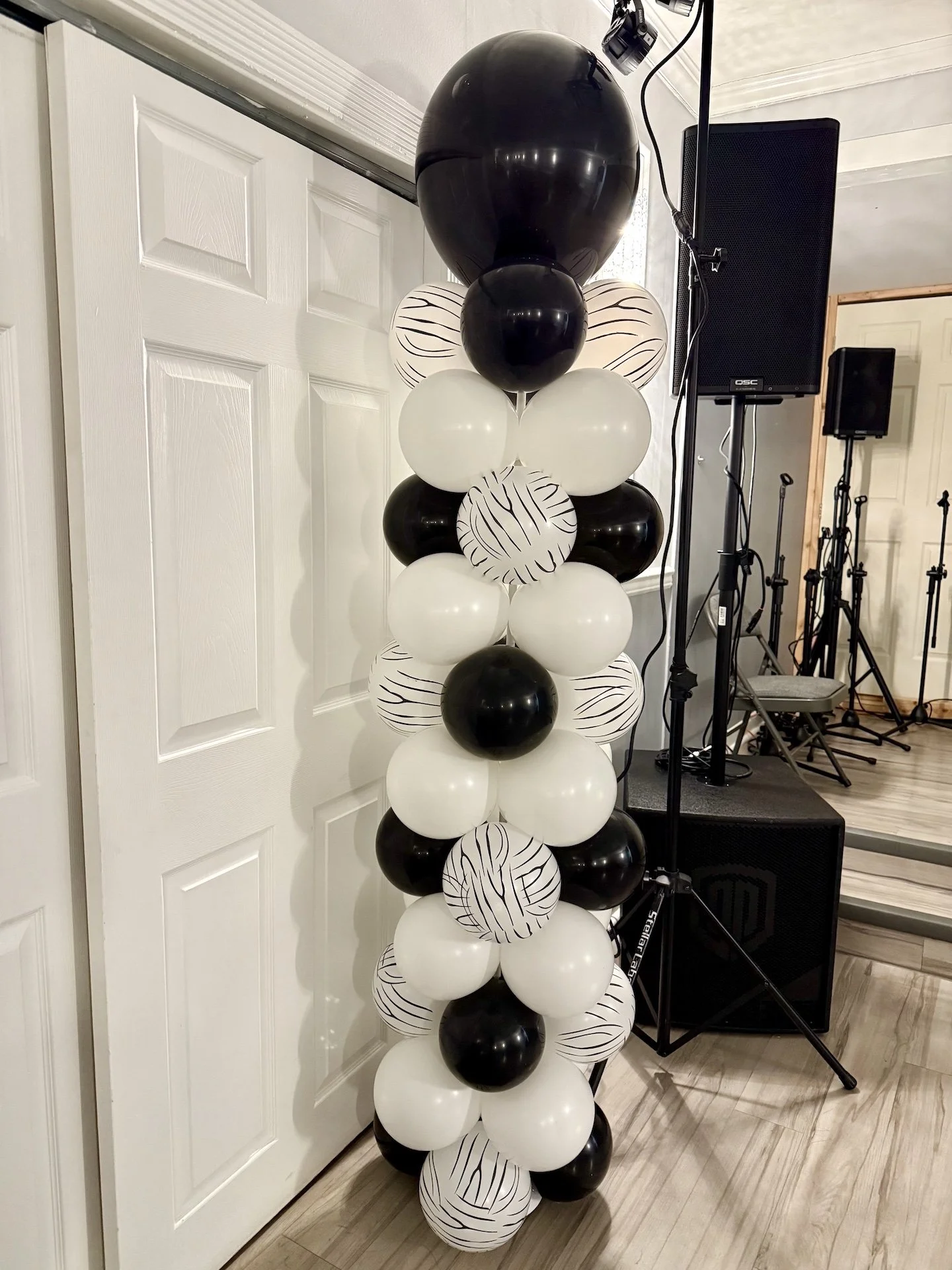 A vertical cluster of black and white balloons, some with striped designs, set up indoors near a white door and audio equipment.