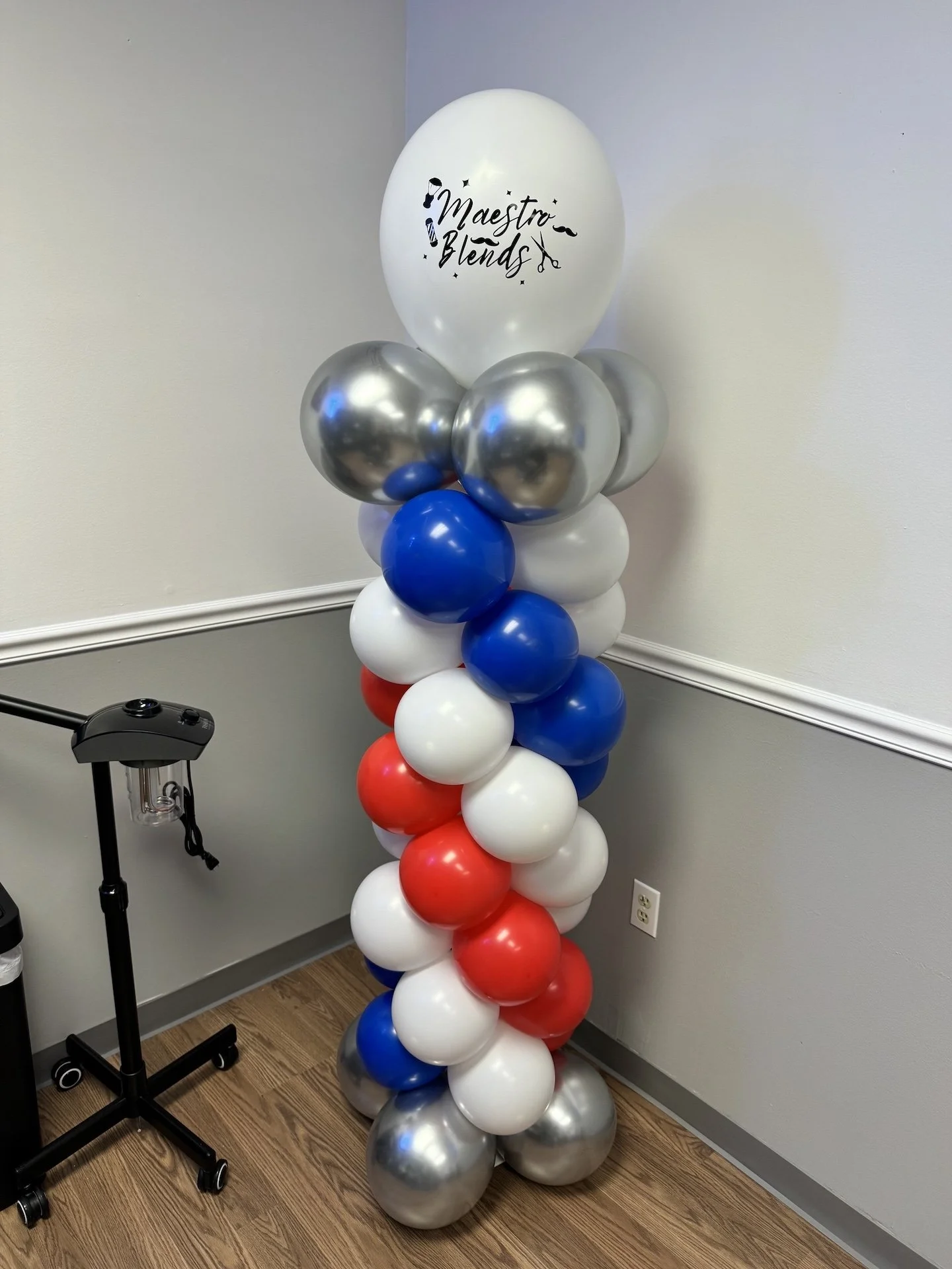 A vertical balloon column with red, white, blue, silver, and white balloons topped with a white balloon that has 'Maestro Blends' written on it, placed in a room corner.