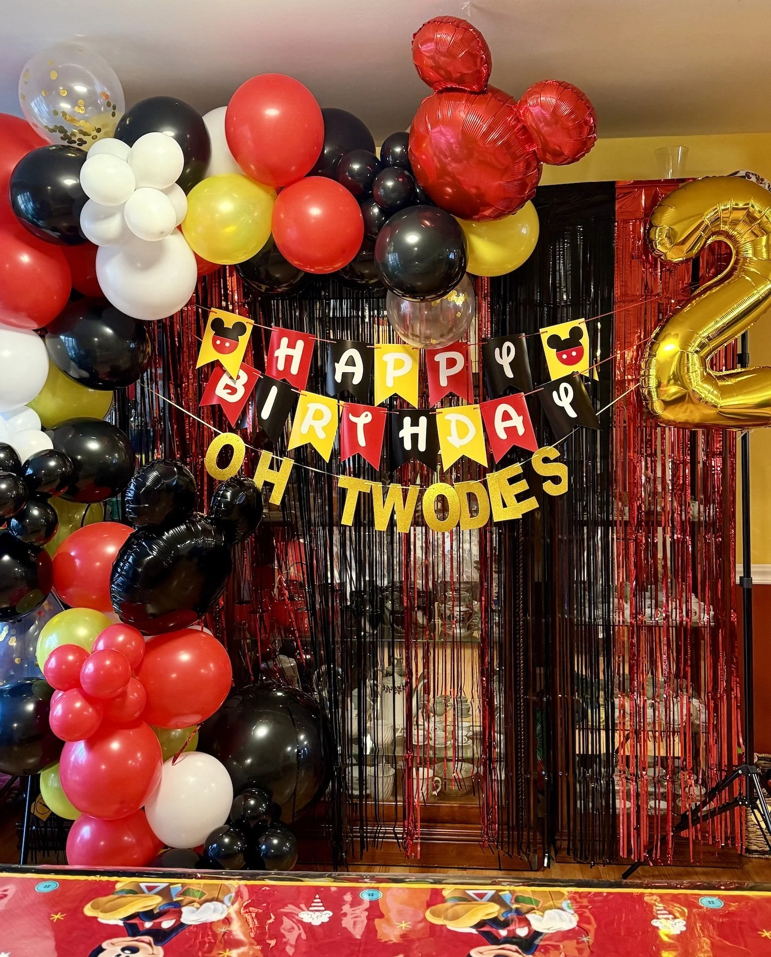 Birthday party decorations with black, red, yellow, and white balloons, a Mickey Mouse themed tablecloth, a "Happy Birthday" banner, and a large number 2 foil balloon.