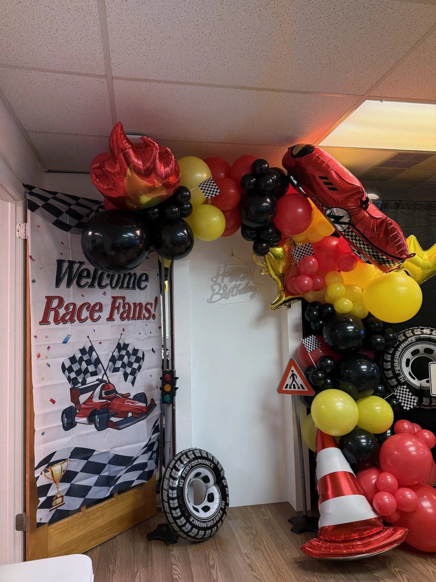 Decorations for a race fan birthday party, featuring a banner with a race car and checkered flags, an assortment of black, red, yellow, and gold balloons, including large race car and tire-shaped balloons, a traffic cone, and a pedestrian crossing si