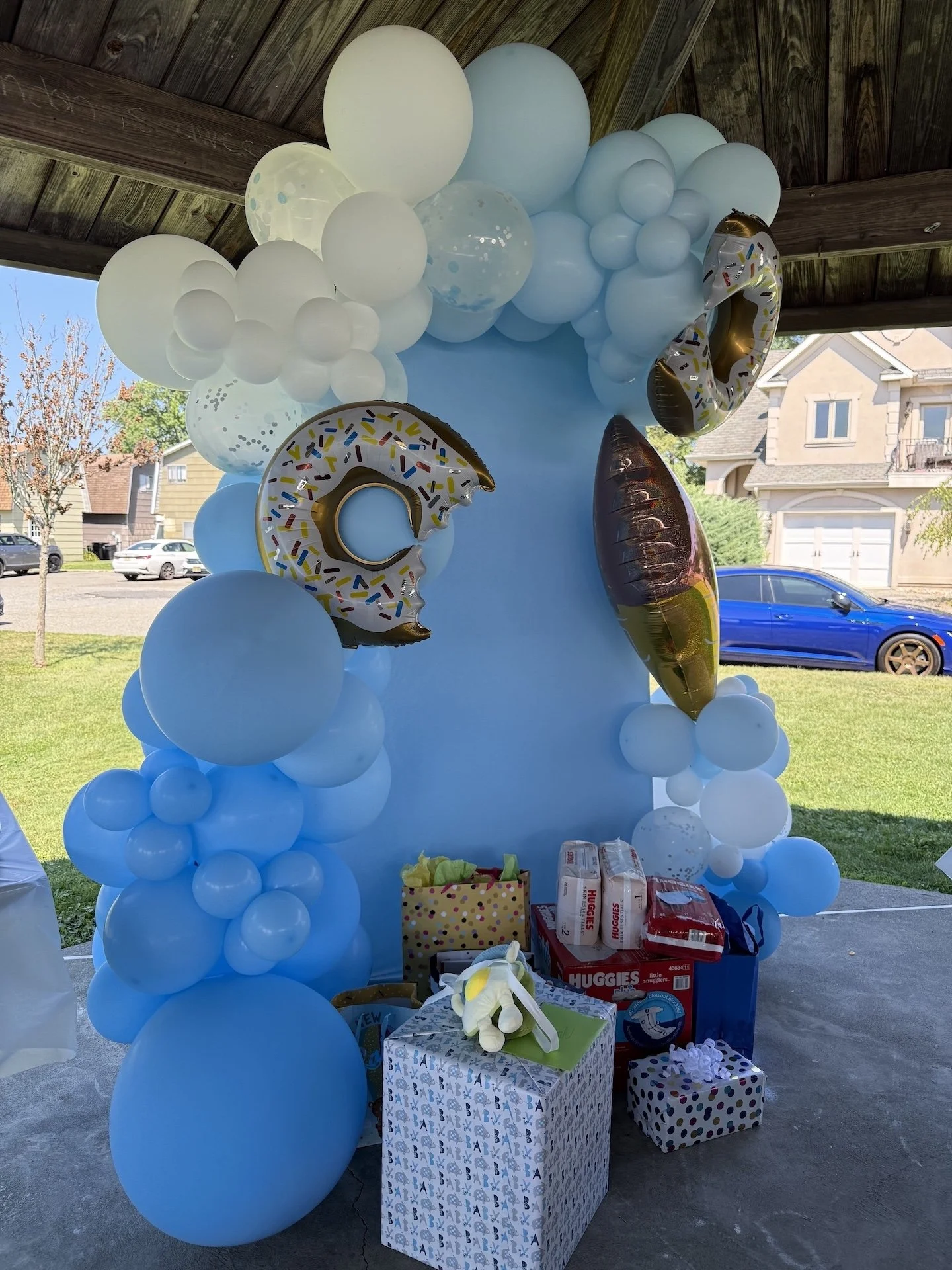 Blue and white balloon arch and decorations with giant donut and ice cream balloon shapes for a baby shower, set outdoors under a wooden shelter with gifts and wrapped presents at the base.