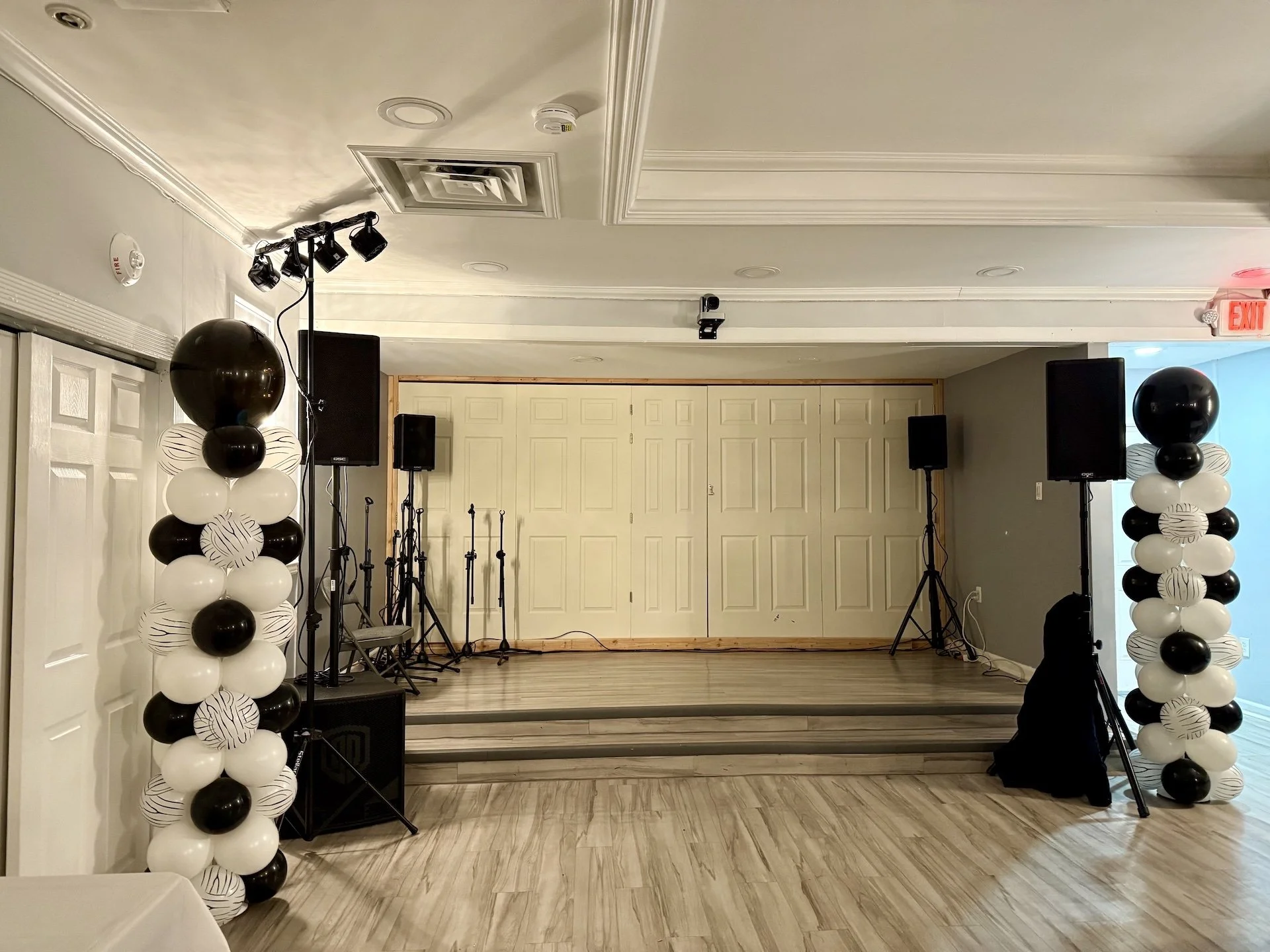 Empty indoor stage with black and white balloon columns on either side, two black speakers, microphones, and lighting equipment, with a closed white double door in the background.