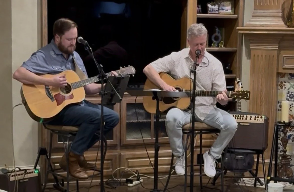 Two men playing acoustic guitars and singing into microphones in a cozy indoor setting.