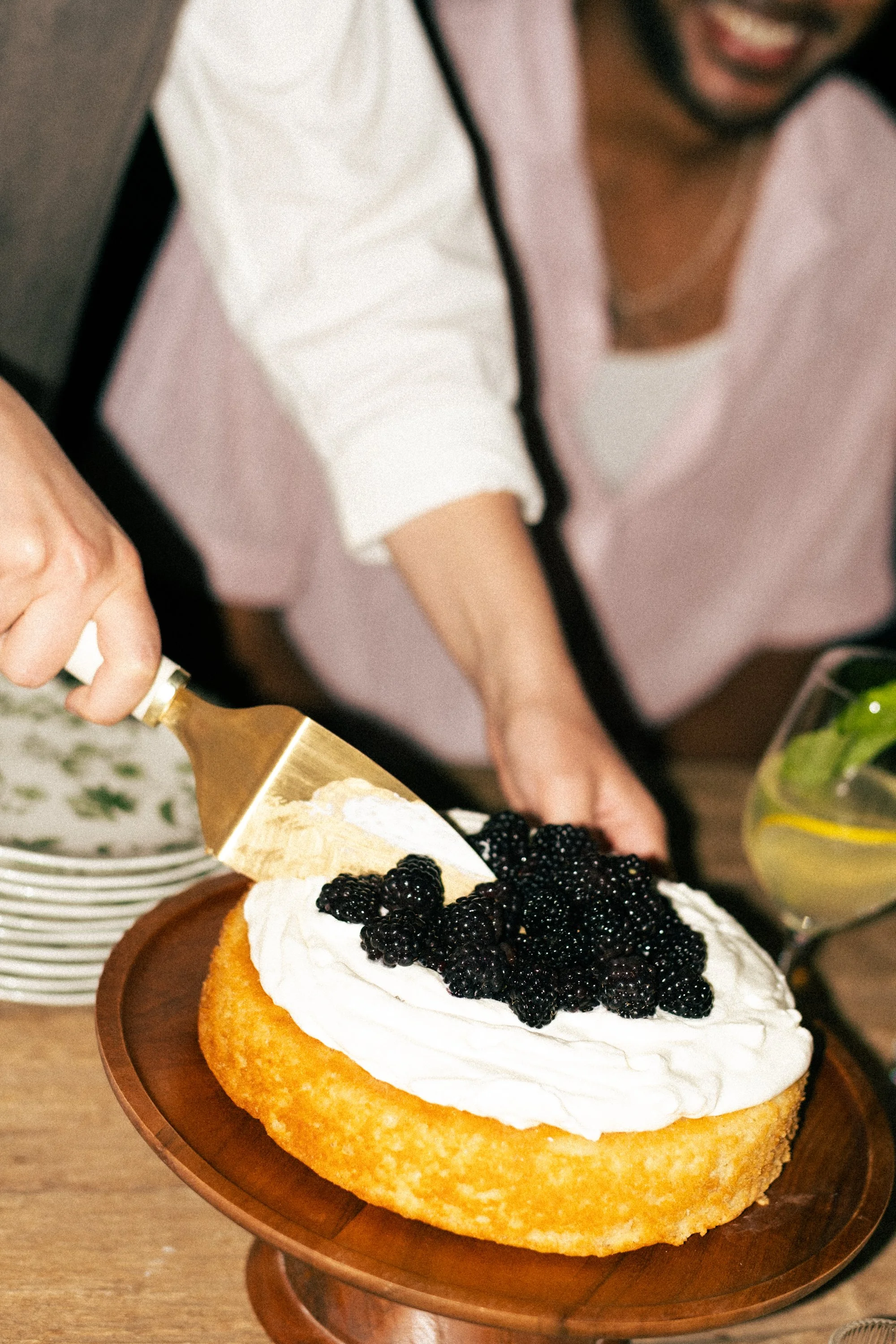 A person is pouring whipped cream over a cake topped with blackberries, placed on a wooden cake stand.