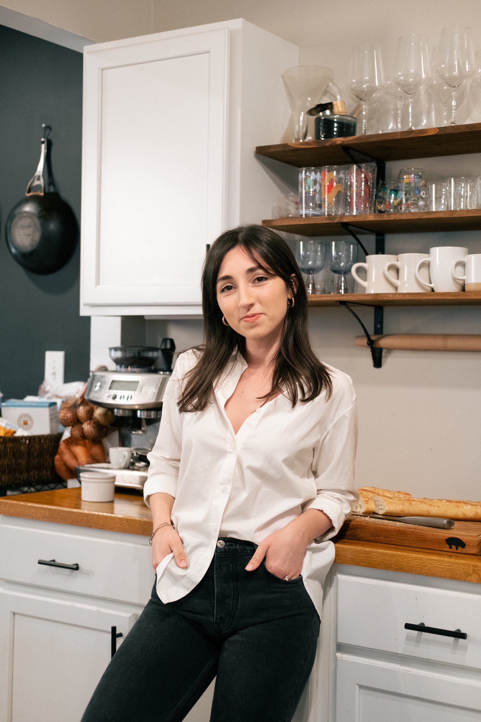A woman with long dark hair and wearing a white button-up shirt and black pants is standing in a kitchen, leaning against a white cabinet with her left hand in her pocket. She is smiling slightly and looking at the camera. The kitchen has open wooden shelves with glassware and mugs, a black frying pan hanging on the dark wall, and a countertop with a coffee machine and some other items.