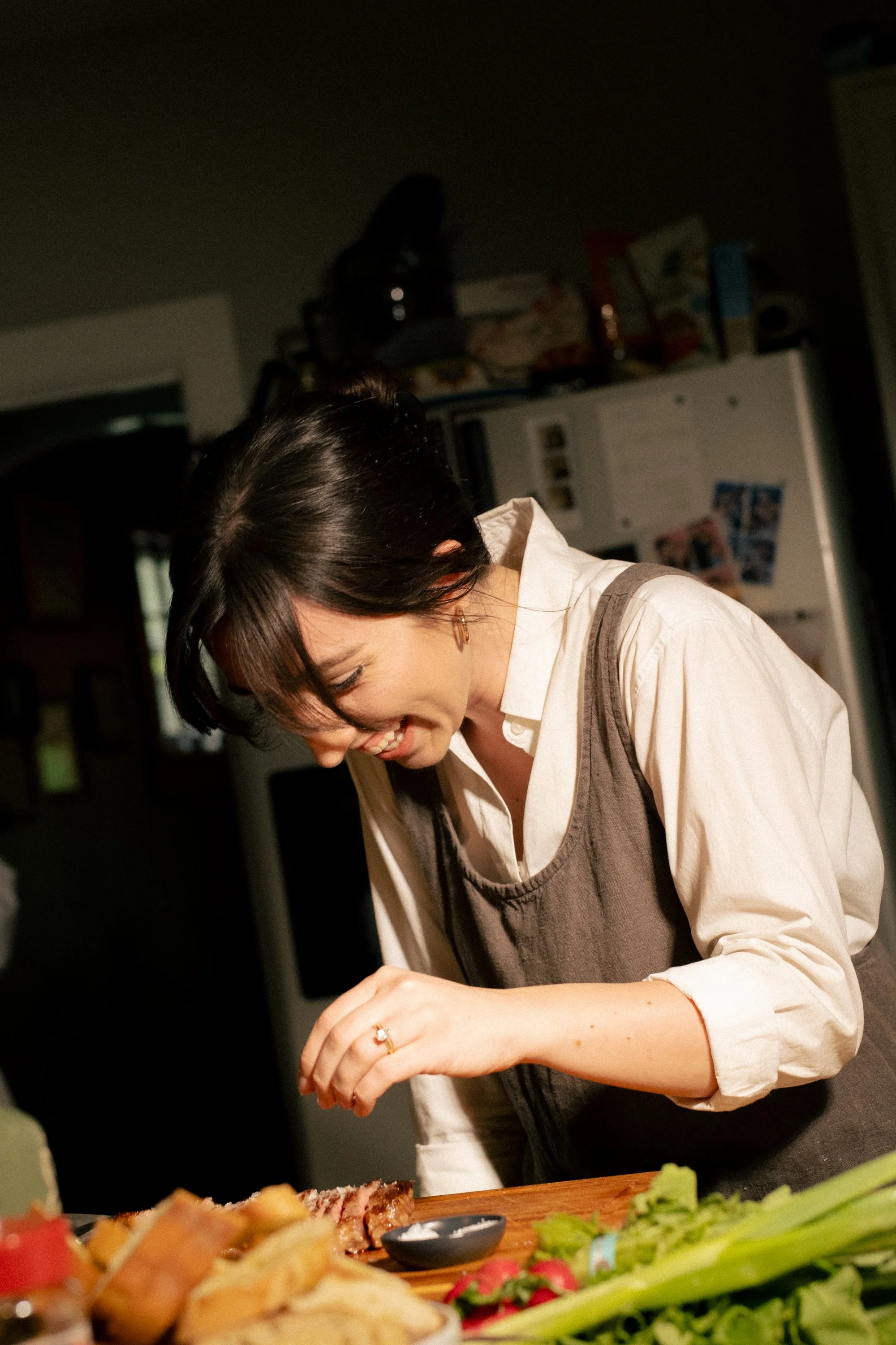 A woman with short dark hair smiling as she prepares food in a kitchen, with a variety of dishes and vegetables on the table in front of her.