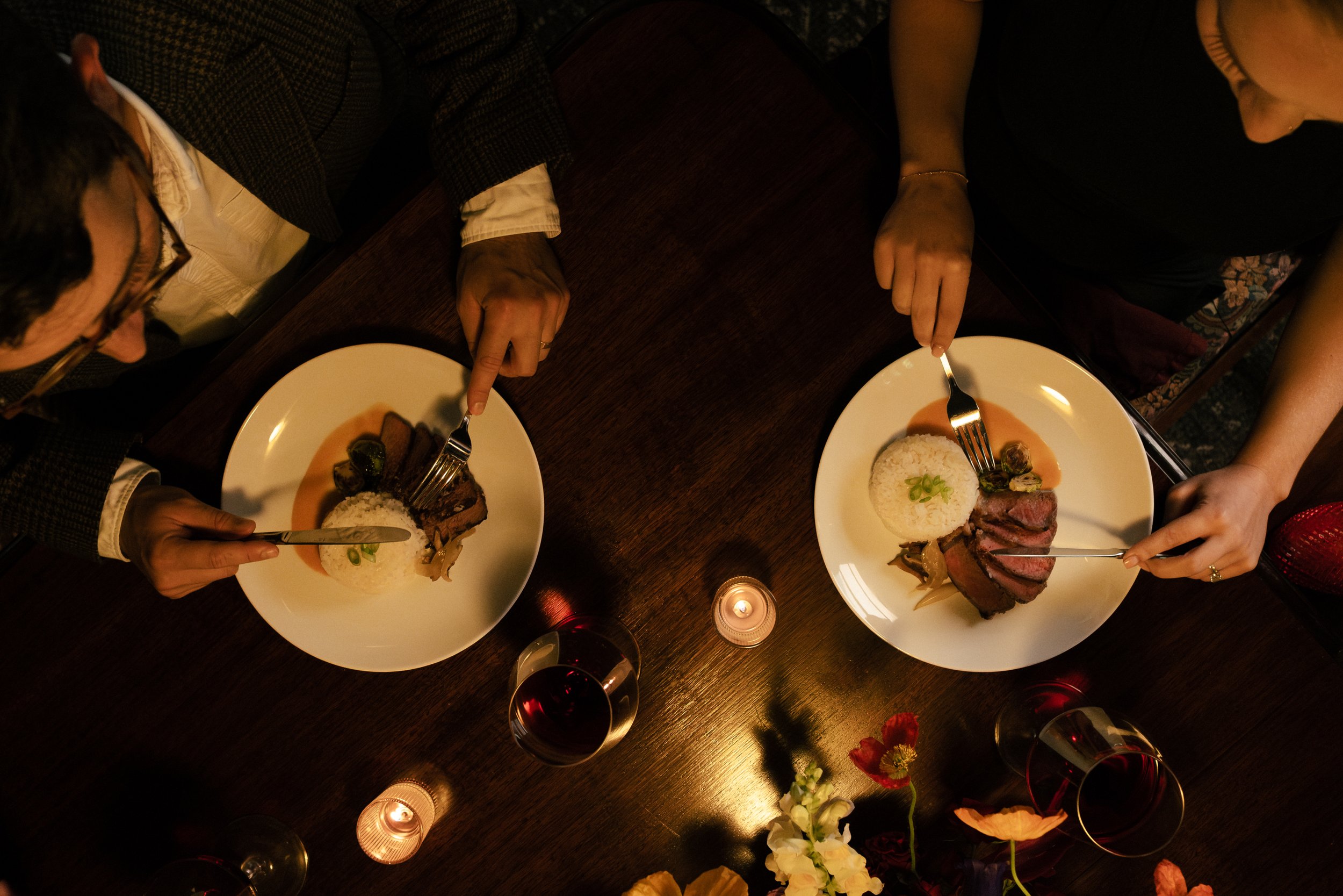 Two people sitting at a dark wooden table with plates of food and glasses of red wine, surrounded by candles and flowers.