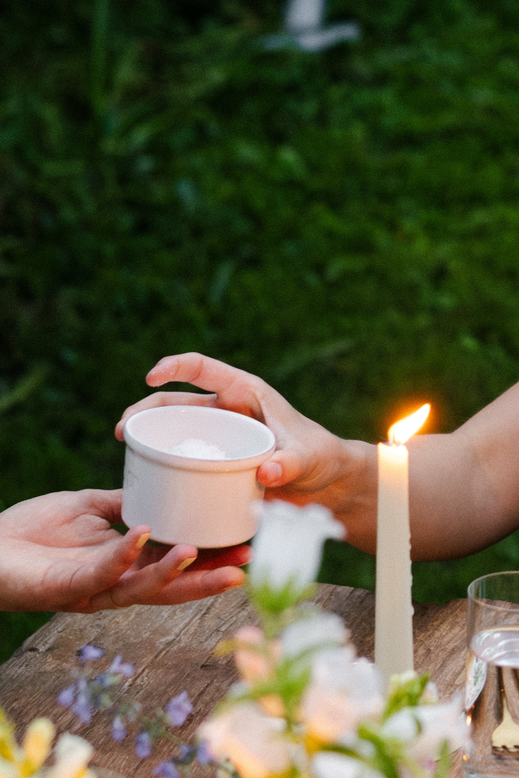 Close-up of hands exchanging a white candle and small white container with a lid, with blurred background showing greenery and a lit candle on a wooden surface.