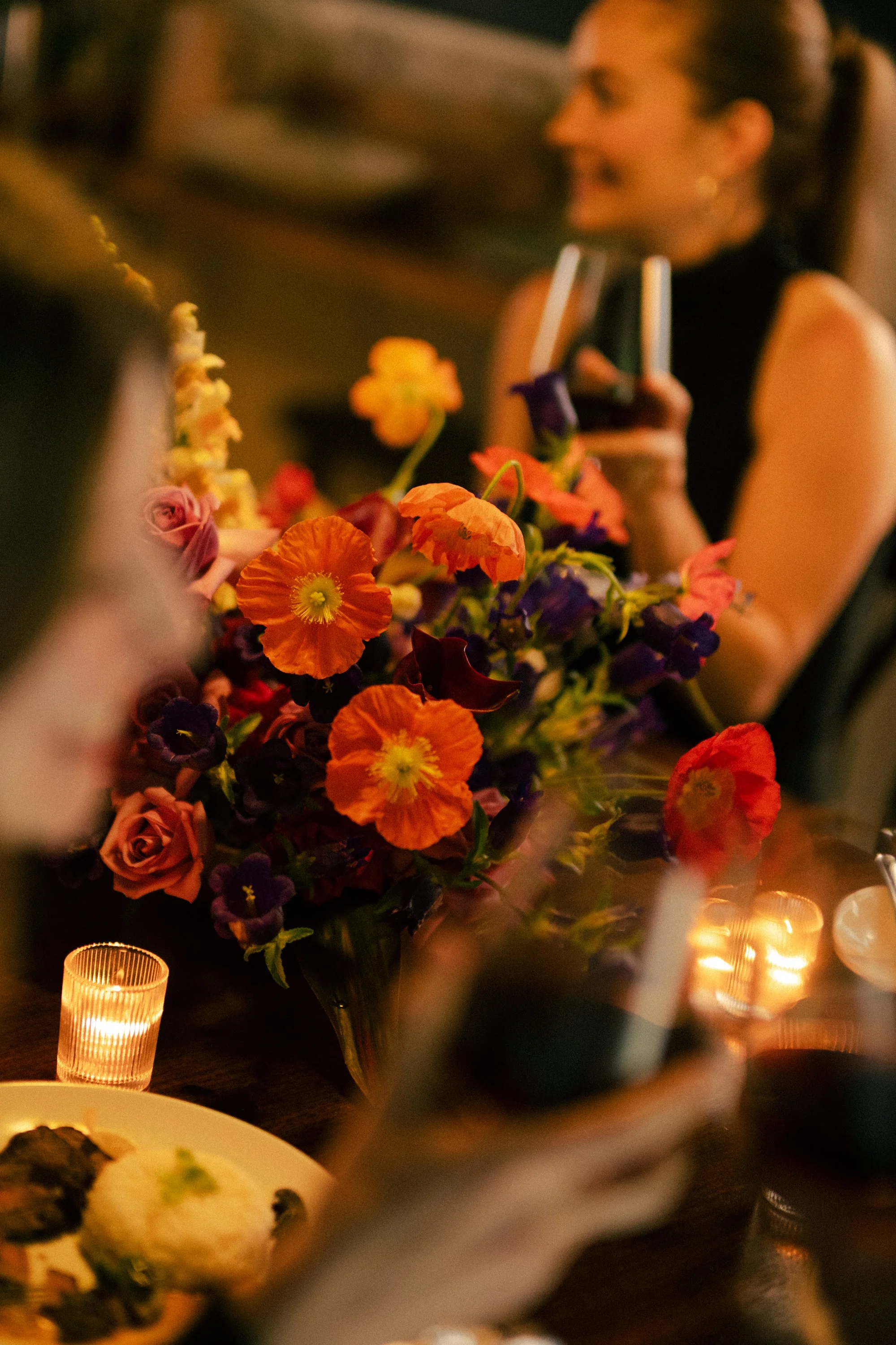 A woman with brown hair in a ponytail smiling, holding a glass, at a dinner gathering with a floral centerpiece and candle lights.