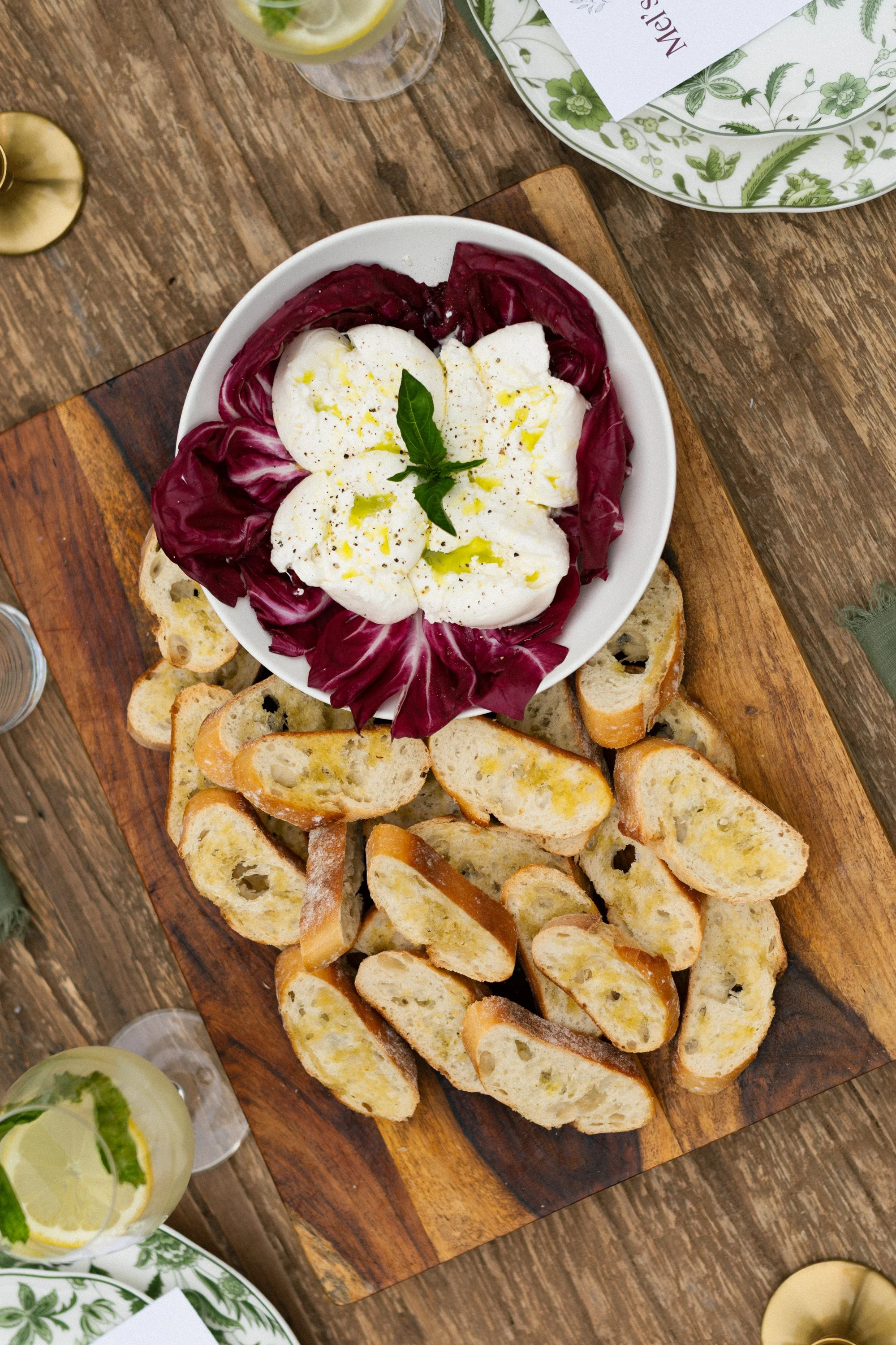 A wooden serving board with sliced toasted bread, a white bowl with burrata cheese garnished with basil and olive oil, accompanied by radicchio lettuce, on a rustic wooden table. Part of a grilled lemon and a glass of lemon water are visible.