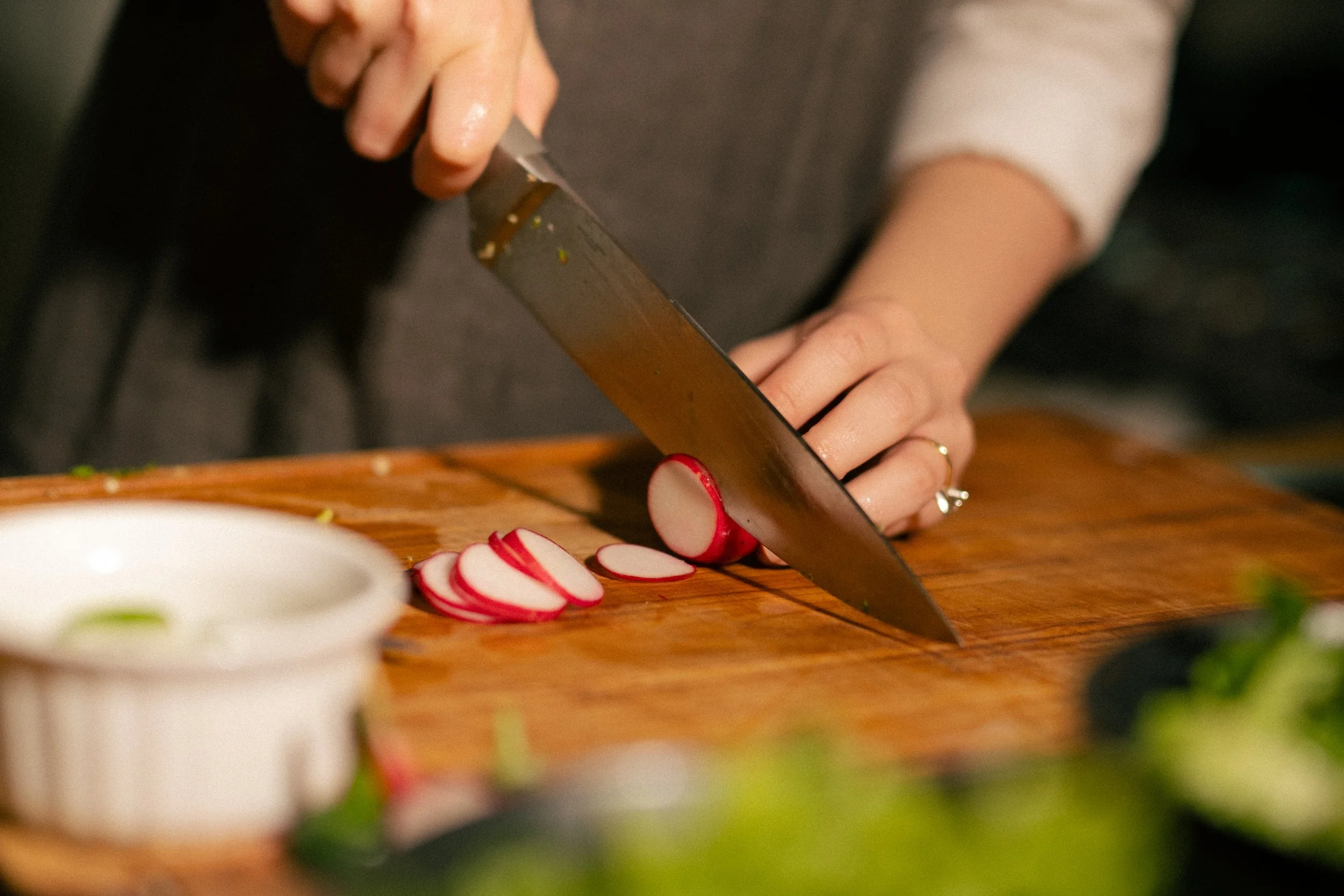 A person slicing radishes on a wooden cutting board with a large kitchen knife.