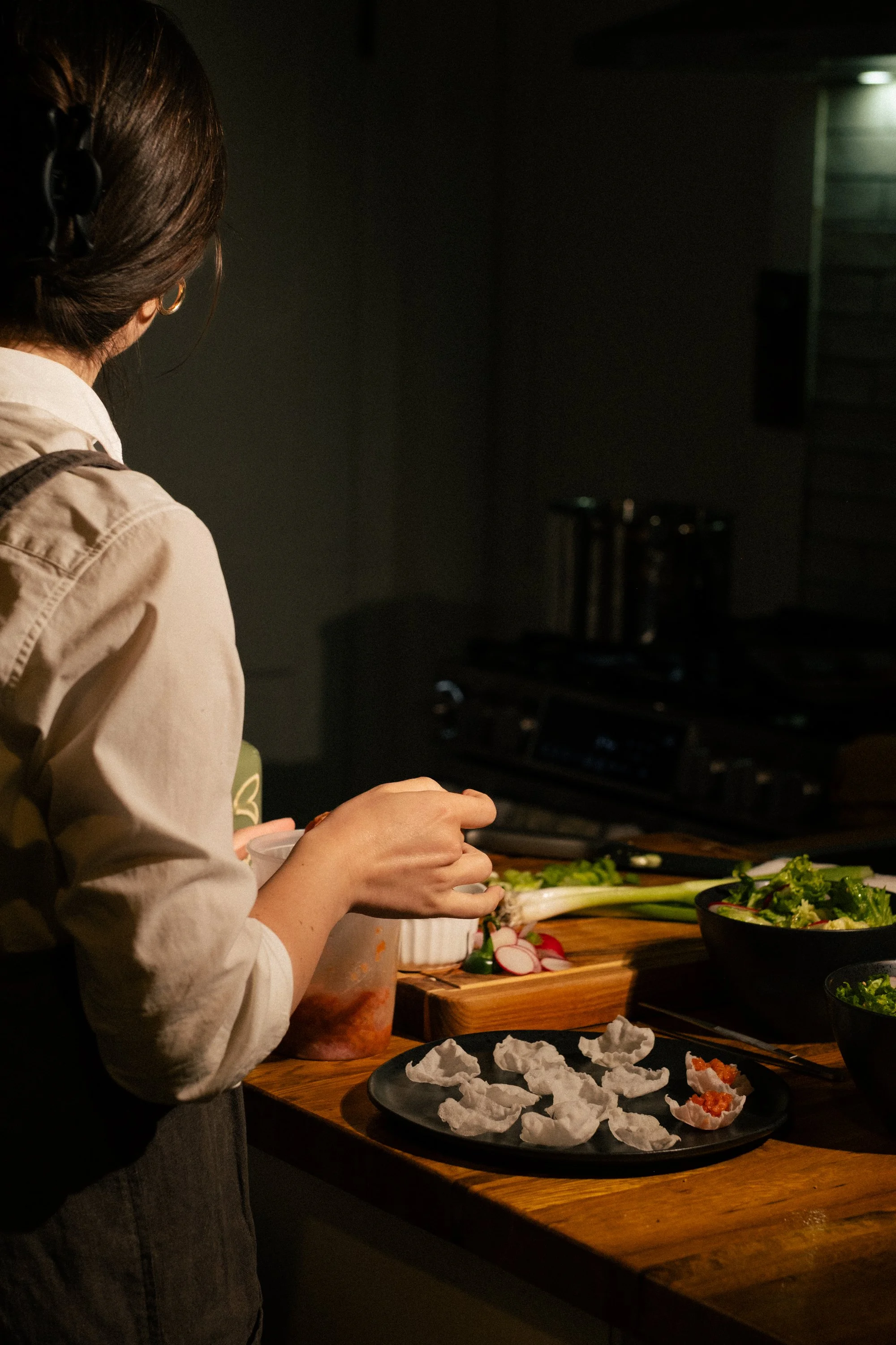 A person preparing food in a dark kitchen, with vegetables and bowls on a wooden countertop.