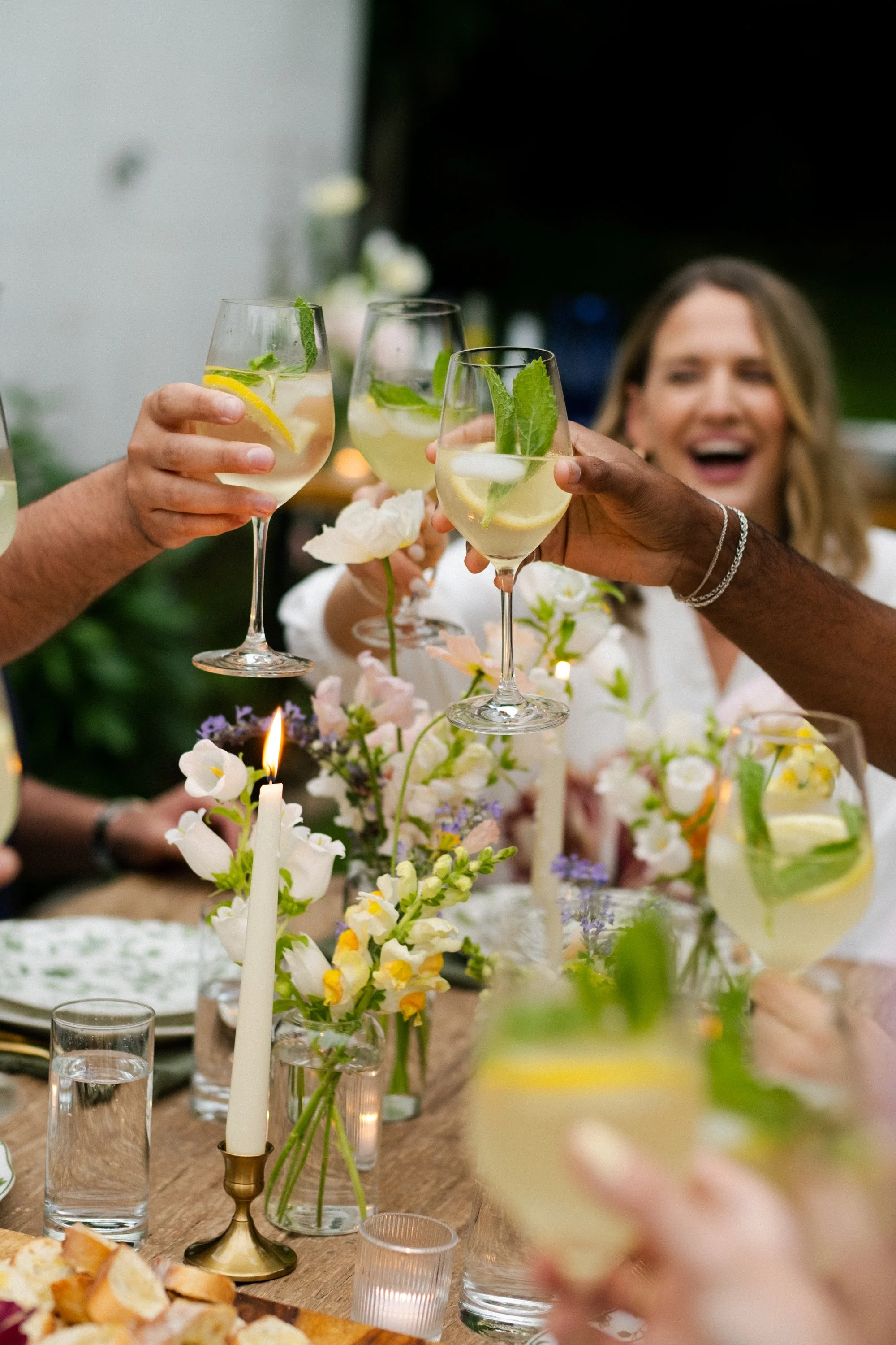 People toasting with glasses of cocktail garnished with mint and lemon slices at a decorated table with flowers and candles.