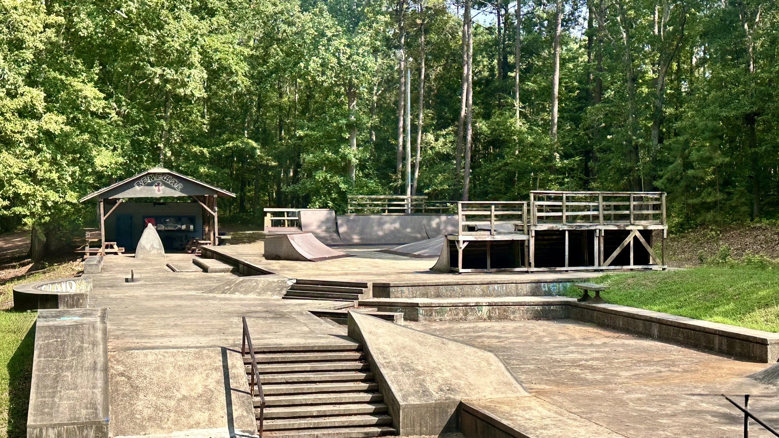 An outdoor skate park with concrete ramps, ledges, and stairs surrounded by green trees and grass.