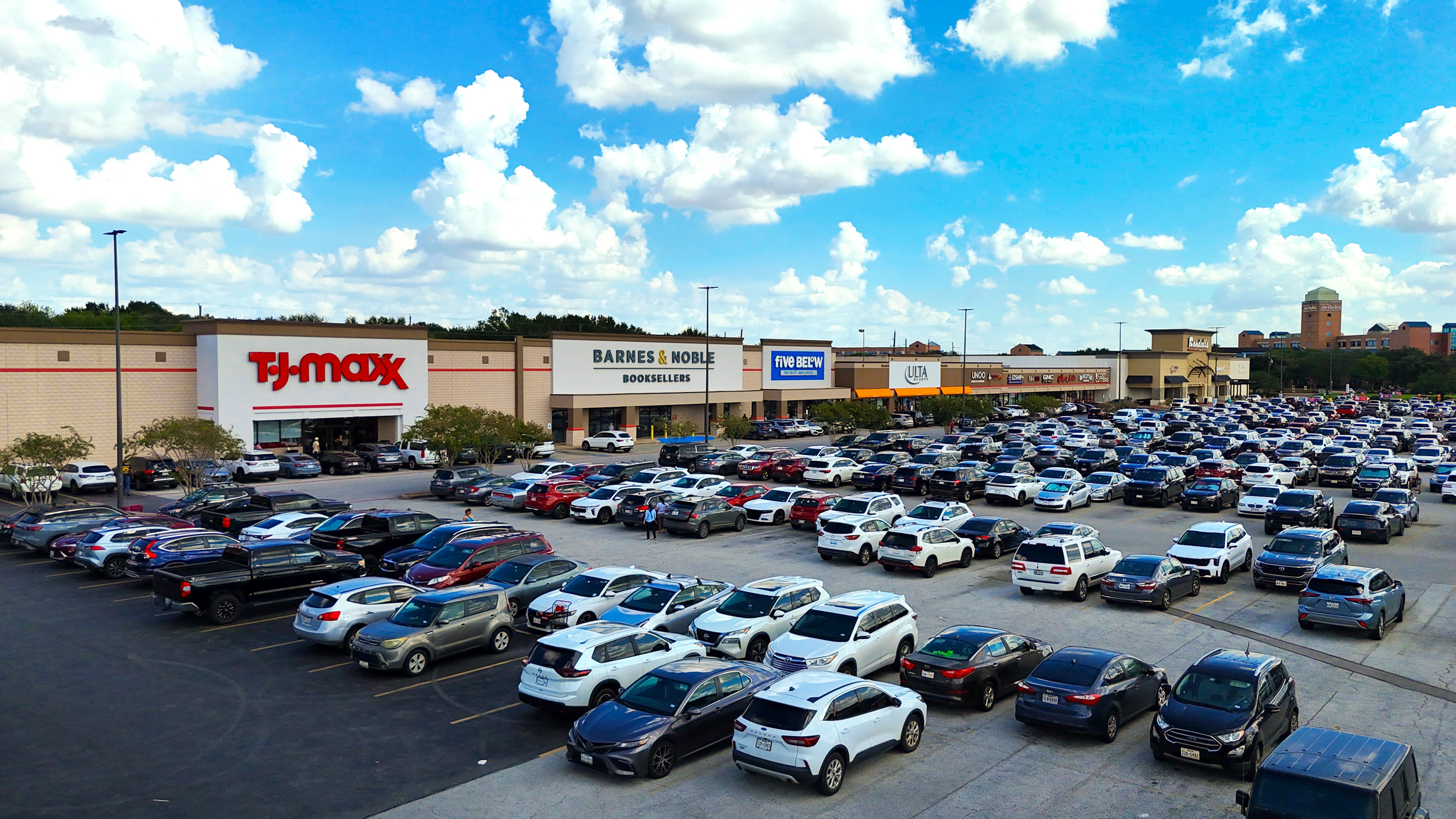 A large parking lot filled with cars in front of a shopping mall. The mall has stores like TJ Maxx, Barnes & Noble, Five Below, and others. The sky is partly cloudy with blue and white clouds.