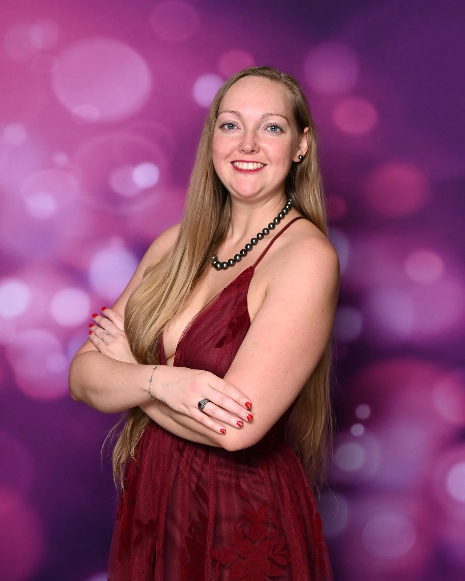A woman with long blonde hair wearing a burgundy dress, black pearl necklace, and red lipstick, standing with her arms crossed against a purple bokeh background.