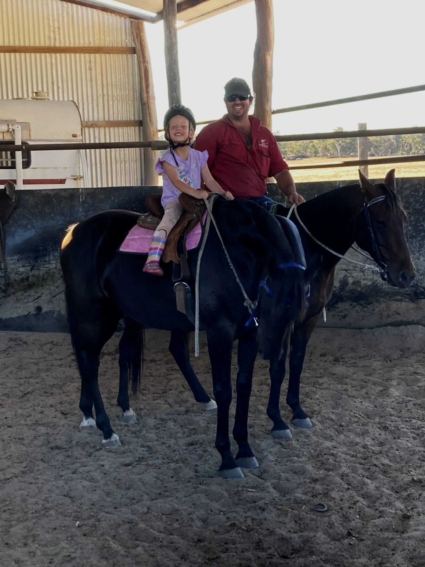 A young girl and a man riding a horse in a covered outdoor riding arena. The girl is smiling and wearing a helmet, while the man is smiling and wearing sunglasses and a red shirt.