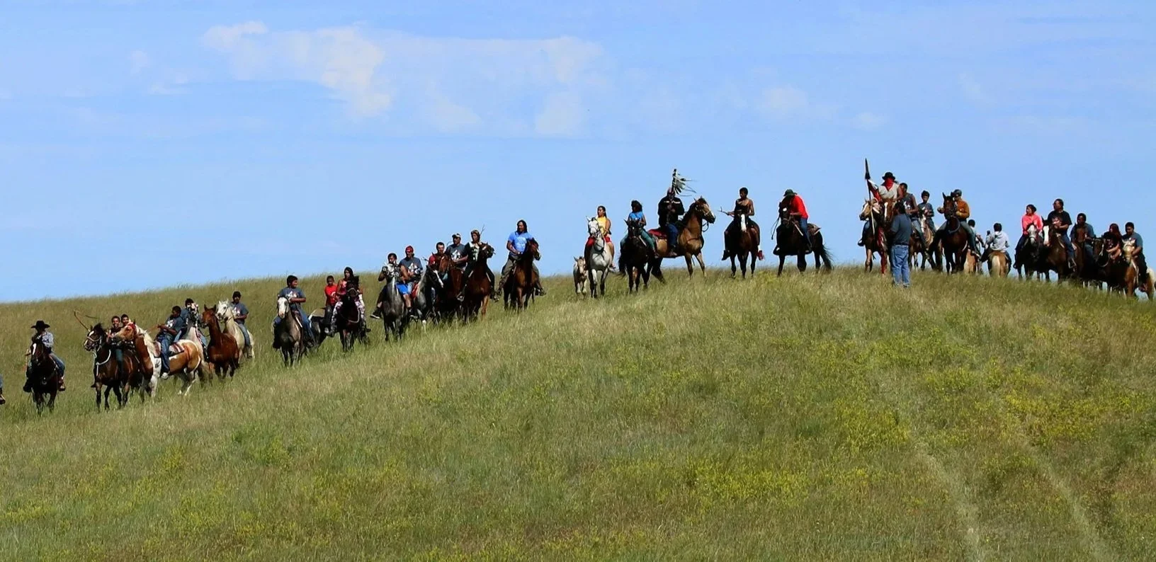 A large group of people riding horses on a grassy hill under a partly cloudy sky.