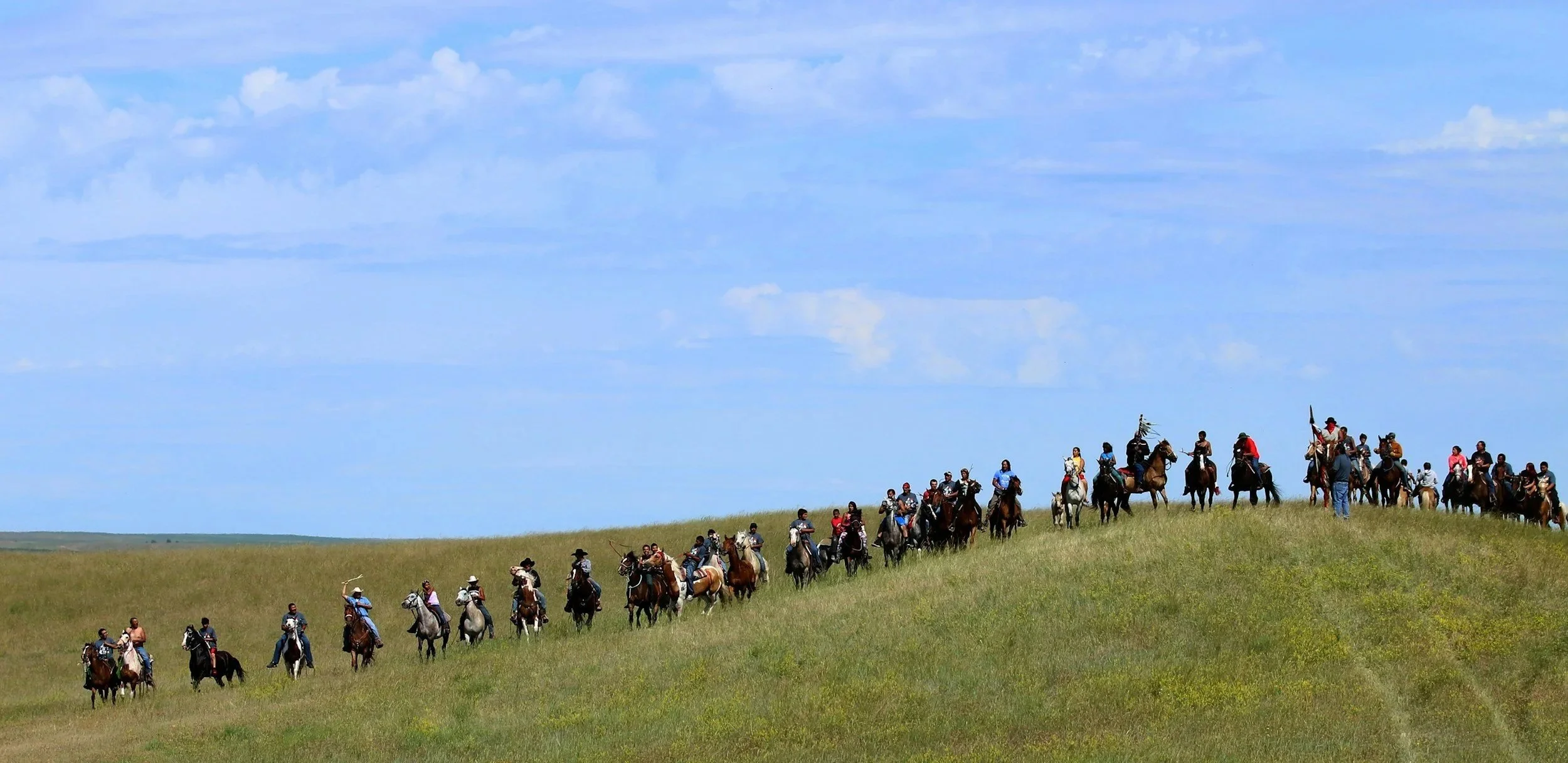 A group of people riding horses across a grassy hill on a clear day with blue sky and clouds.