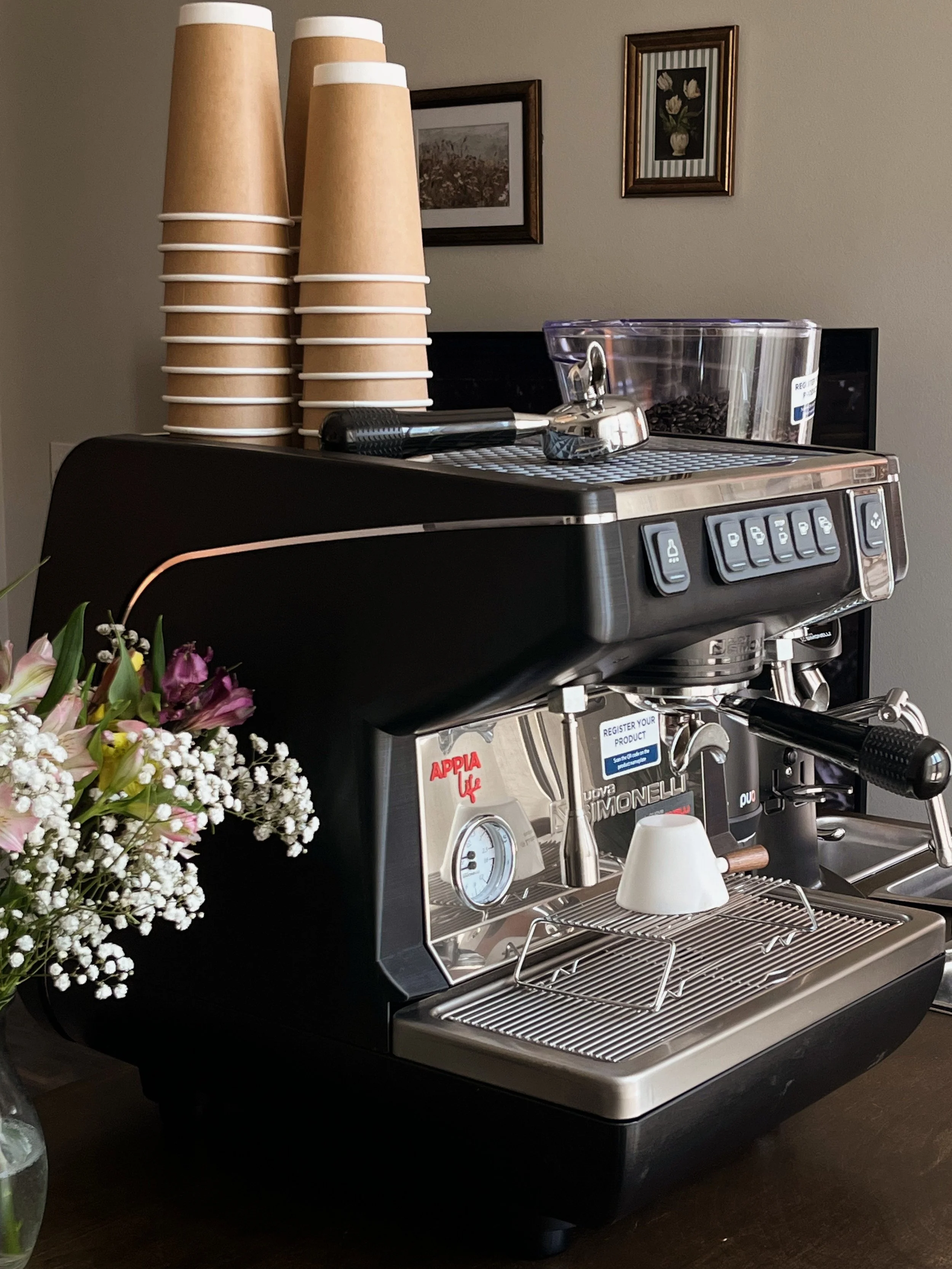 A professional espresso machine with a steam wand, on a dark wooden surface, with a flower arrangement in a vase to the left, and a stack of disposable cups, a tamper, and a grinder with locally roasted espresso beans on top.