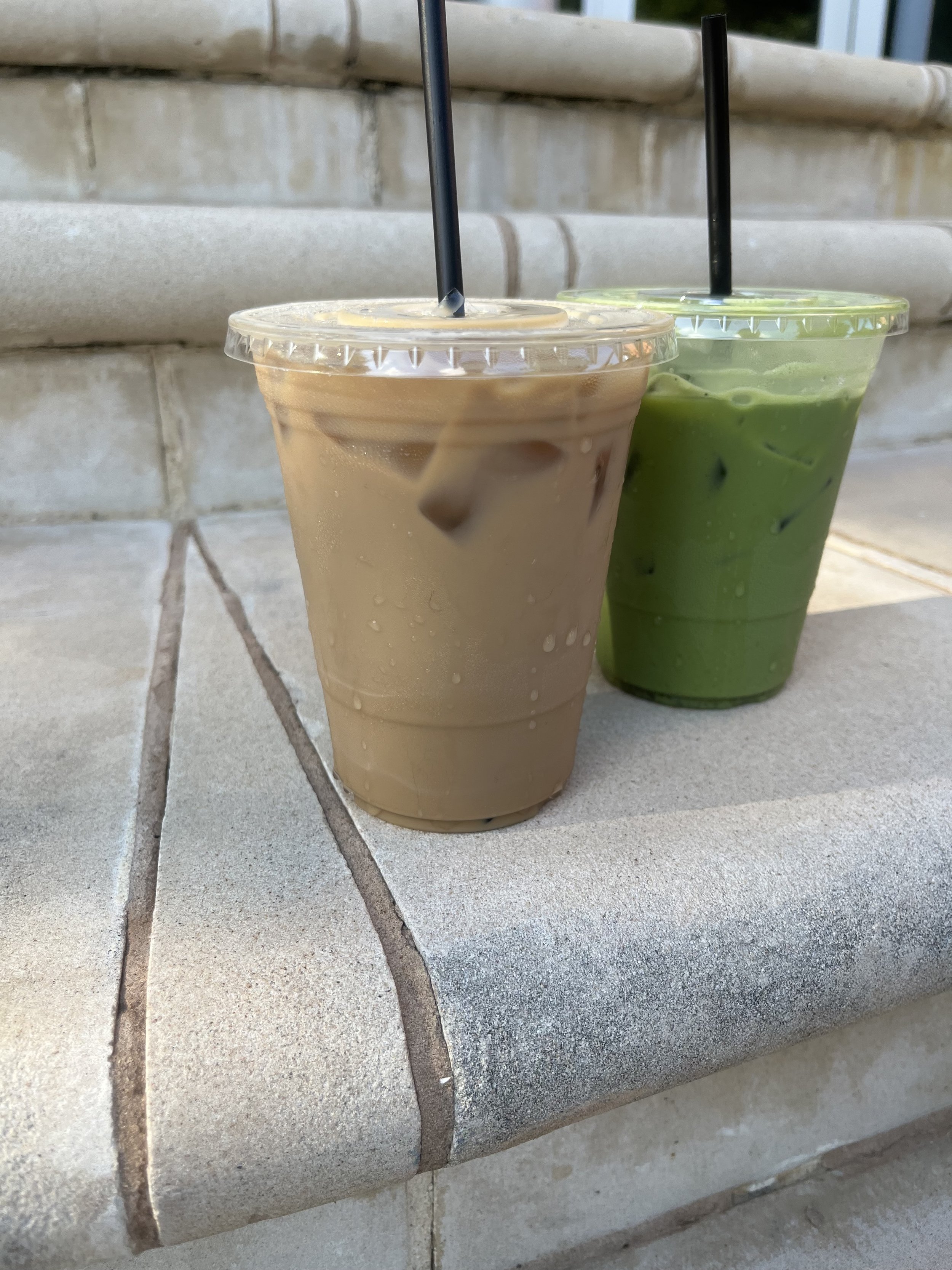 Two iced drinks in plastic cups with black straws sit on a tiled surface in front of a brick wall.