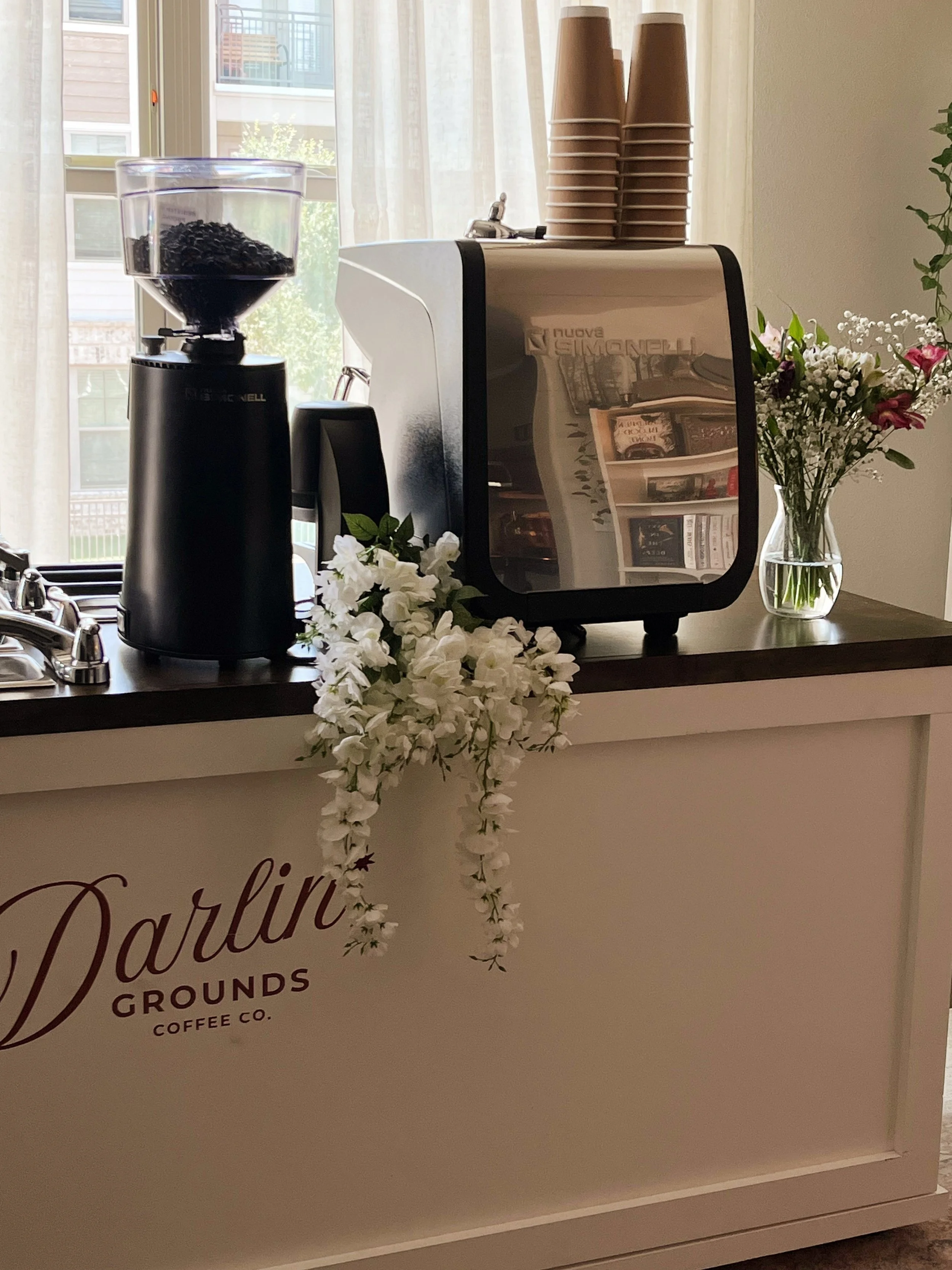 A mobile coffee cart with a coffee grinder, an espresso machine, stacks of disposable cups, and a vase of flowers, with a window in the background.