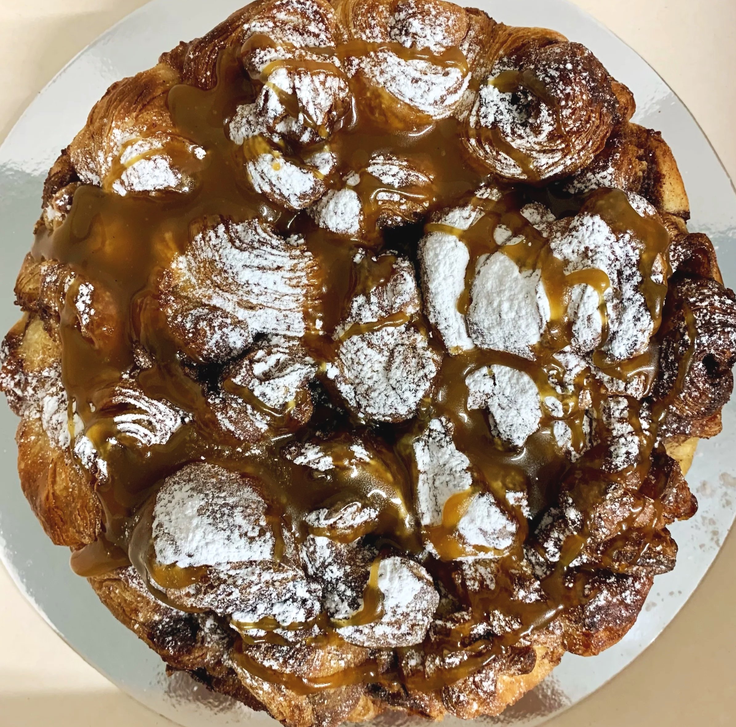A round cinnamon scroll croissant pull apart pastry topped with powdered sugar and caramel sauce.