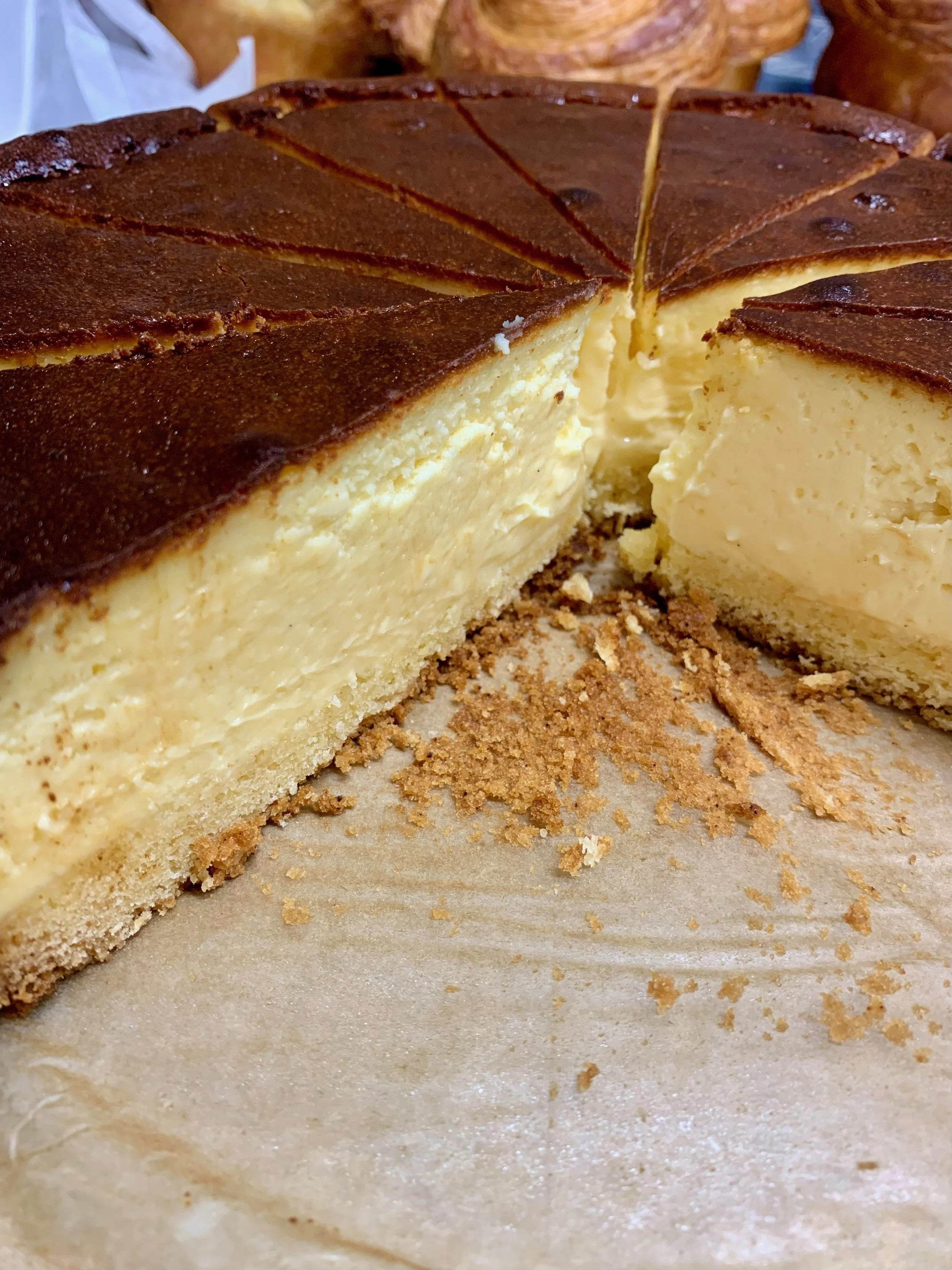 A cheesecake with a graham cracker crust and a caramelized top, partially sliced to reveal a creamy, dense interior, on parchment paper with crumbs scattered around.