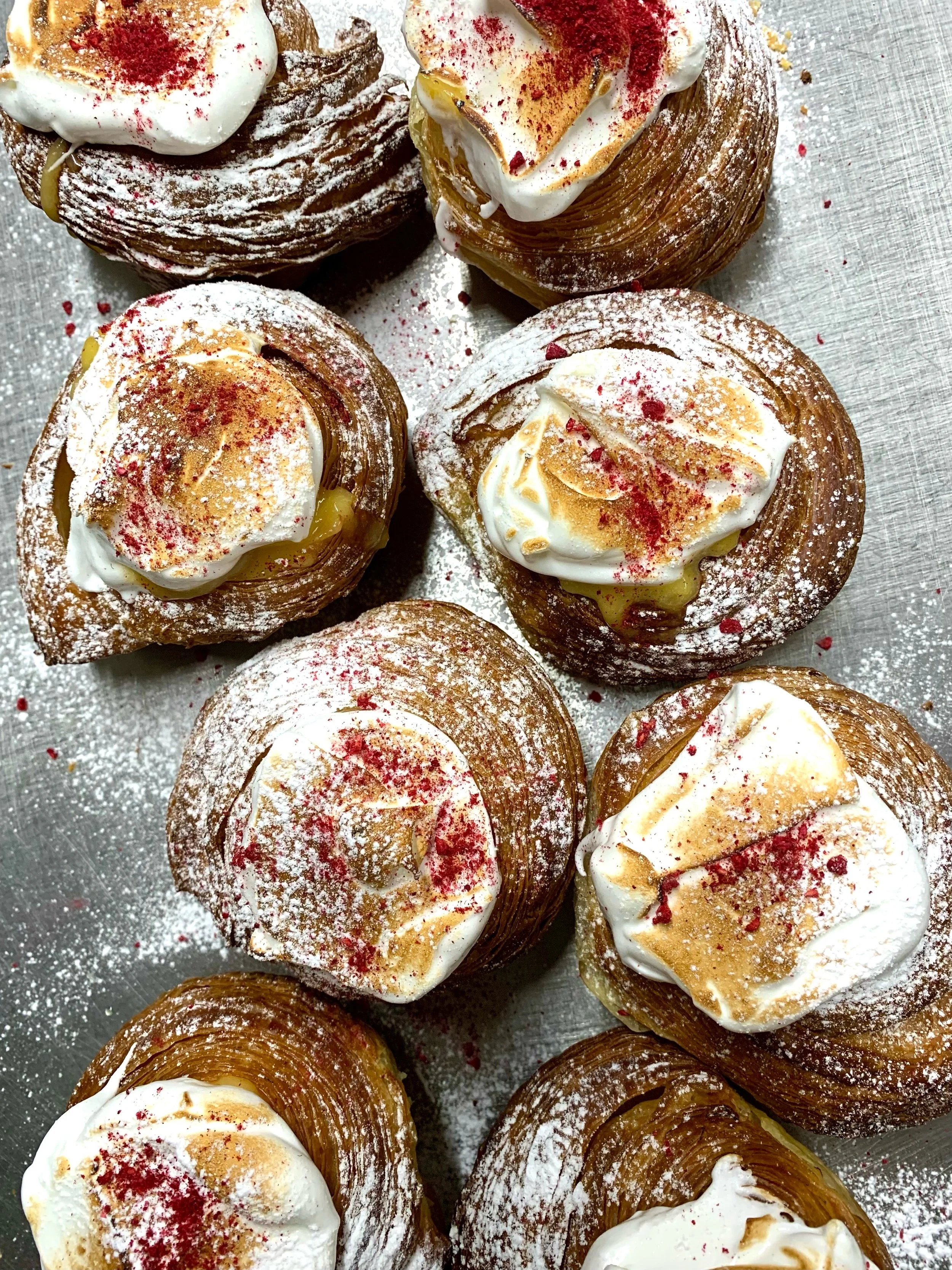 Close-up of several cream-filled pastries dusted with powdered sugar and garnished with red sprinkles, on a metallic surface.