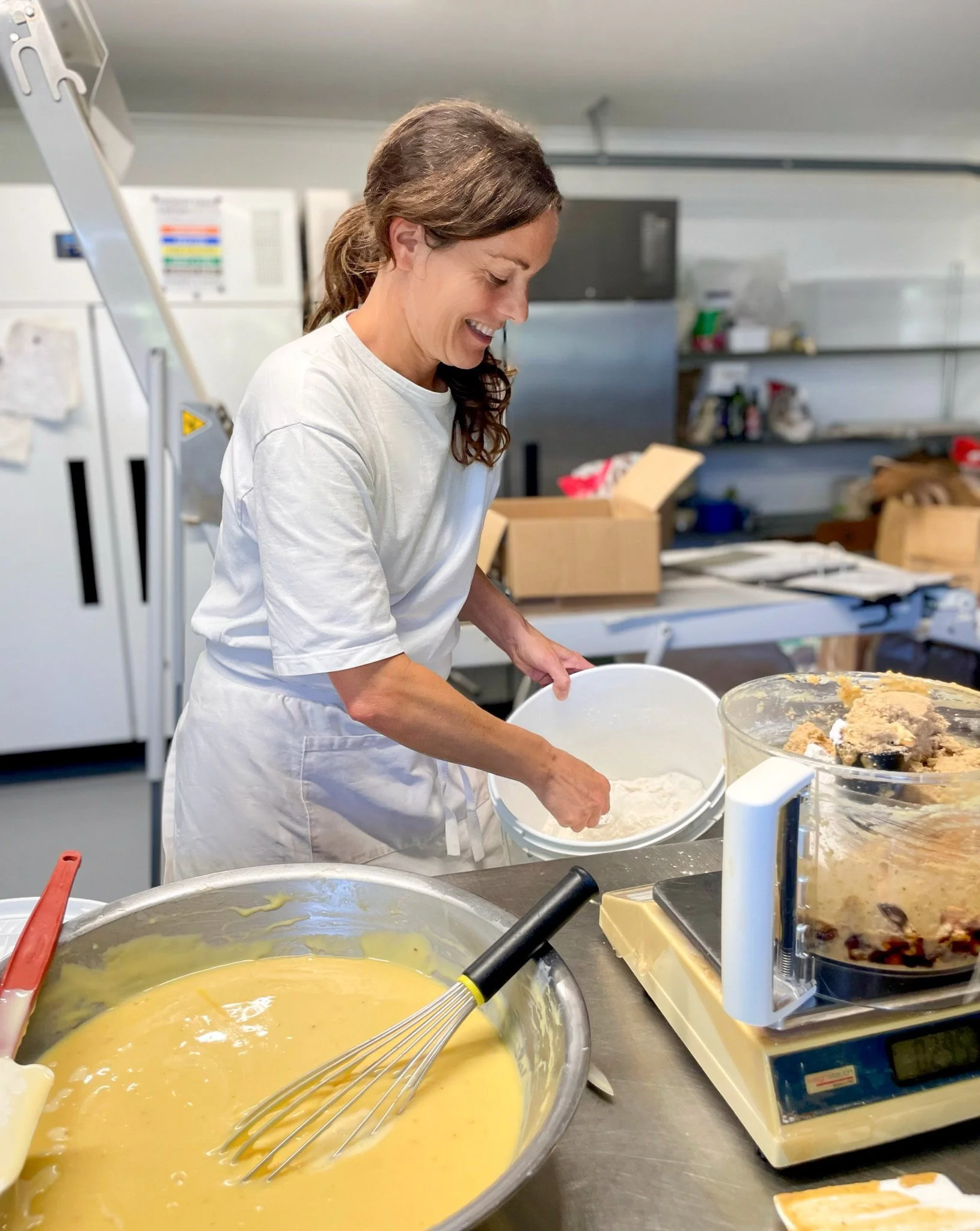 Jennifer Jane Perry owner of Jen's Pastries Mangawhai  wearing a white shirt and apron smiling while baking food in her kitchen, mixing ingredients in a bowl with a large whisk, with various kitchen tools and ingredients on the counter.
