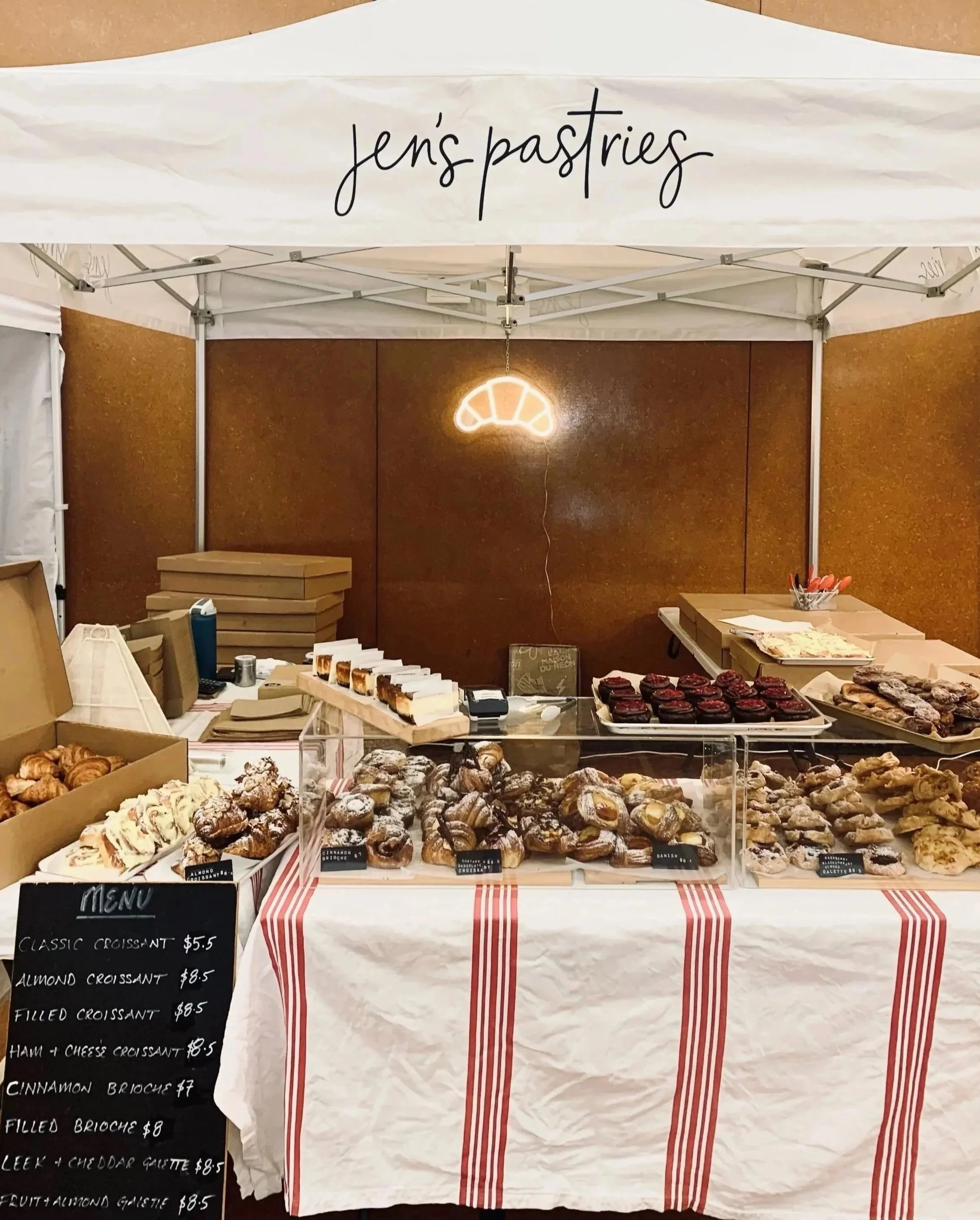 A tiny bakery stall named 'Jen’s Pastries' at Mangawhai Markets with a variety of croissants, pastries, and baked goods on display, a menu board listing different types of croissants and their prices, and a neon croissant sign hanging behind.