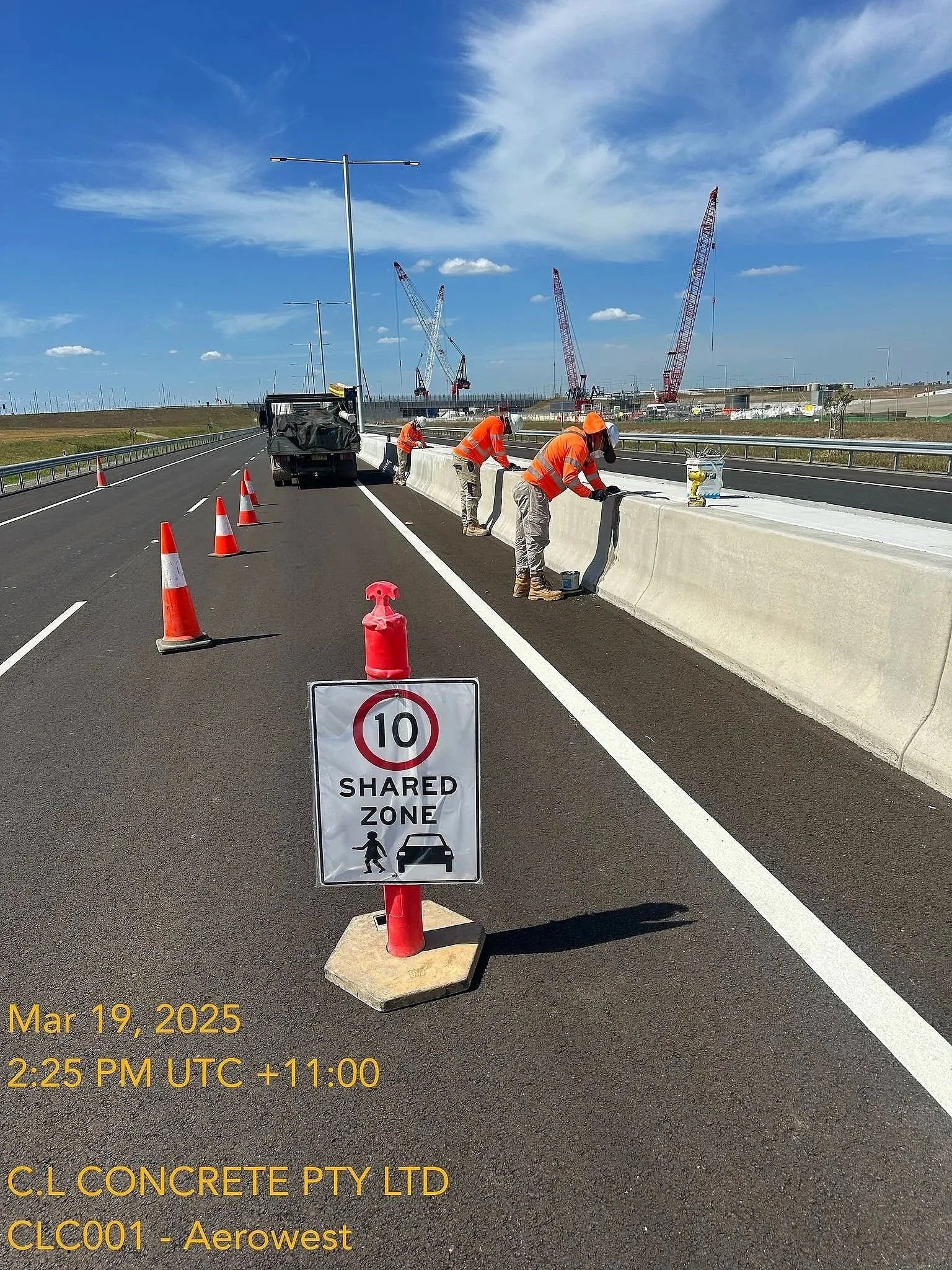 Workers in orange vests and helmets laying concrete barriers on a highway under a blue sky with clouds. Traffic cones and a shared zone sign are visible in the foreground, with cranes and construction equipment in the background.