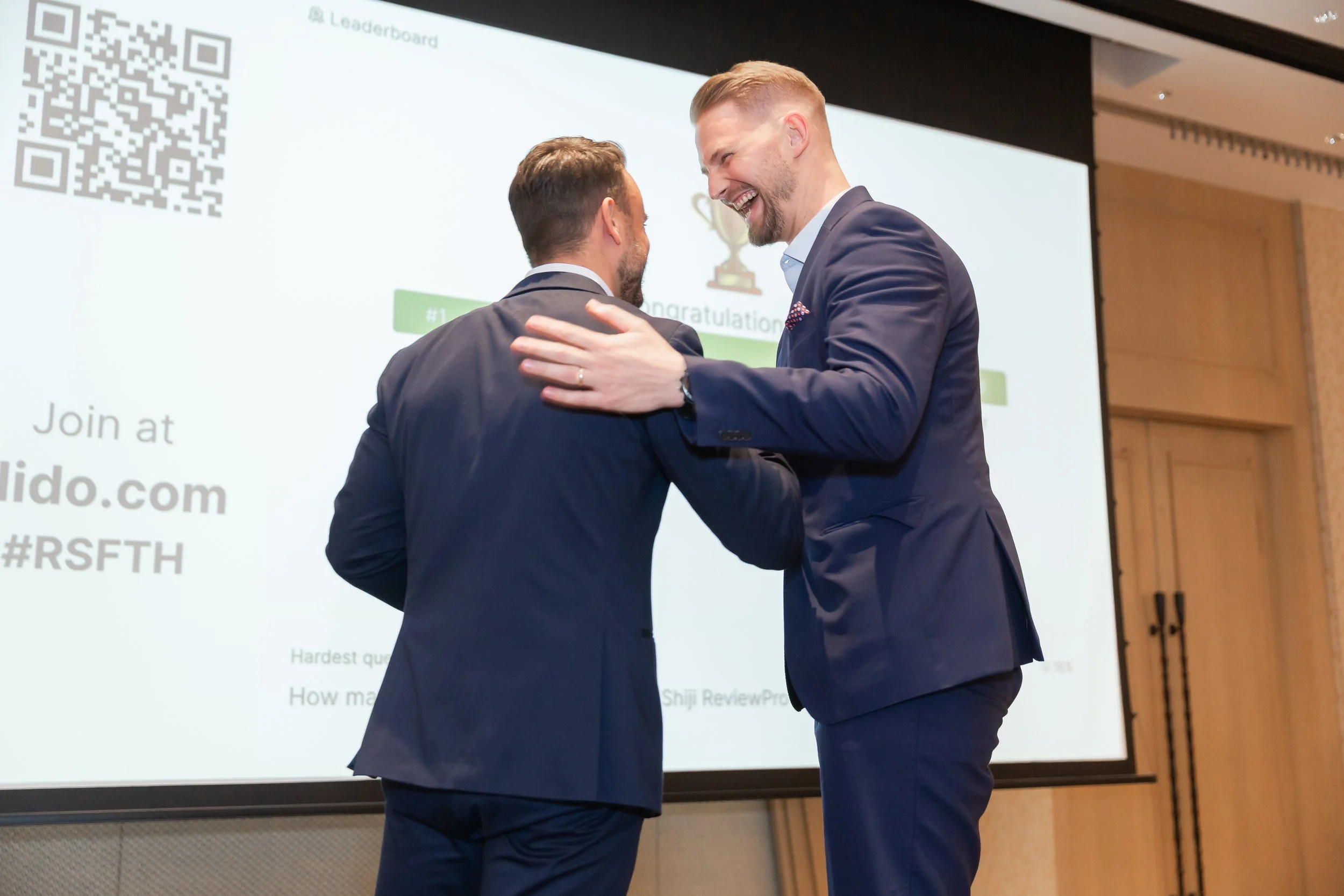 Florian Ultsch and a business partner in business suits smiling and embracing at a conference in Southeast Asia, with a large screen displaying congratulatory text and a QR code in the background.