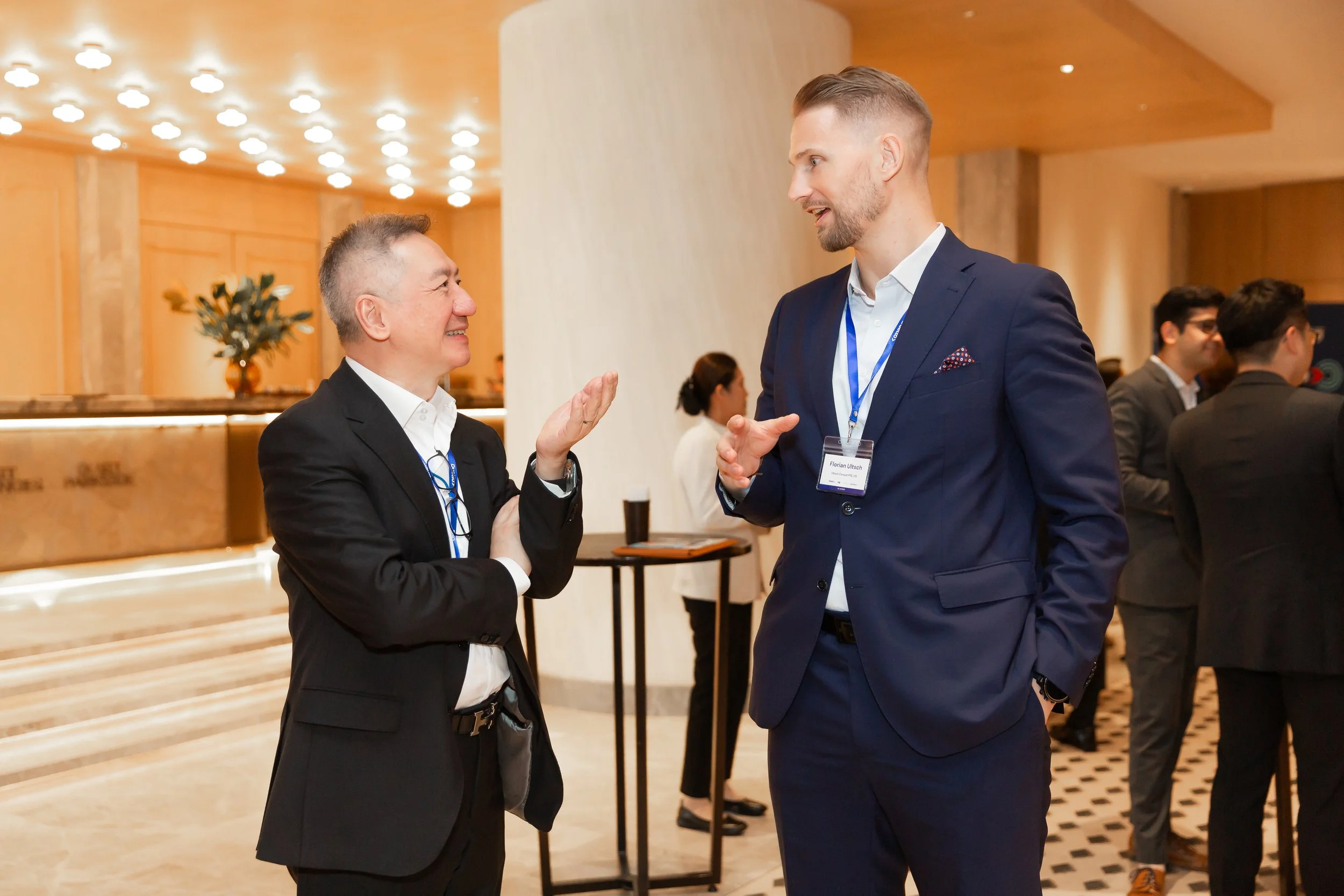 Florian Ultsch and a business partner in suits having a conversation at a professional networking event in Bangkok in a well-lit, modern venue. One man is smiling with arms crossed, while the other gestures with his hand.