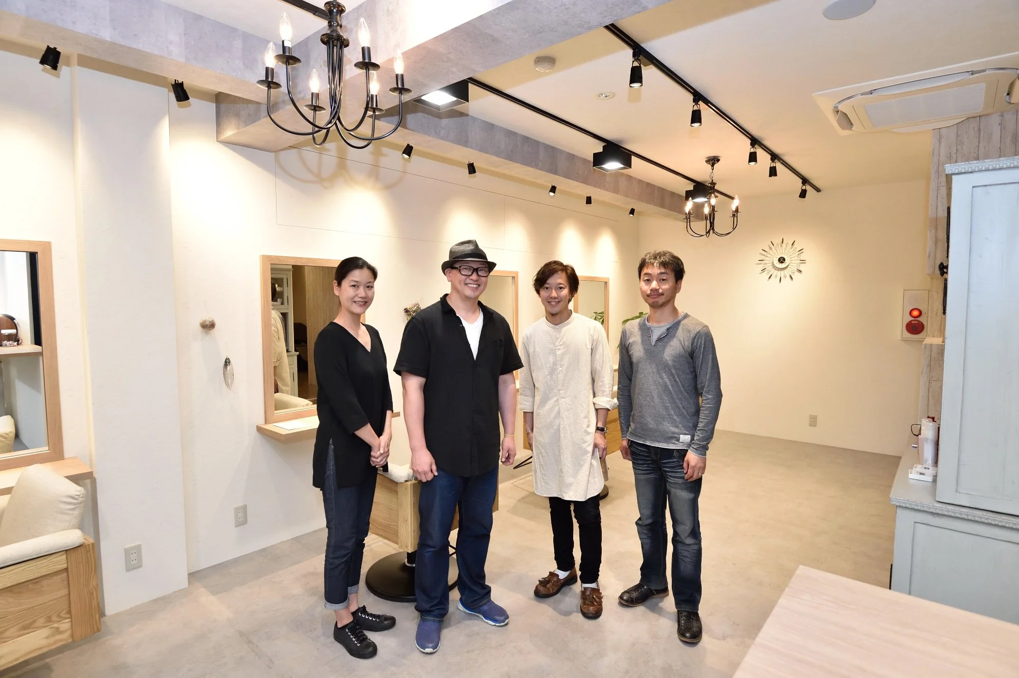 室内で4人の男女が笑顔で並んでいる。背景には照明やミラー、壁に掛け時計が見える。