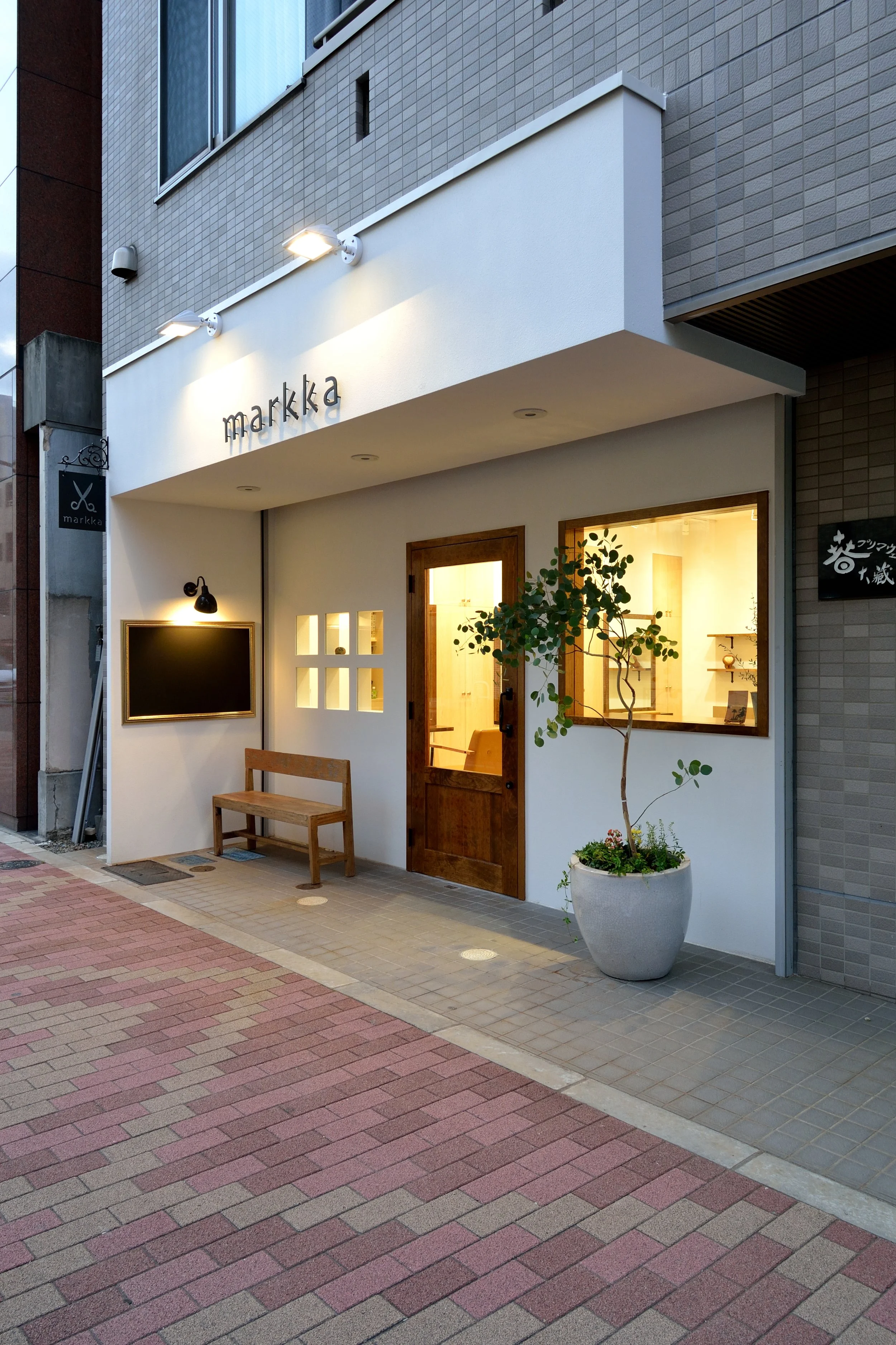 白い建物の外観、木製のドアと窓、緑の植物が植えられた鉢、看板に「markka」と書かれている