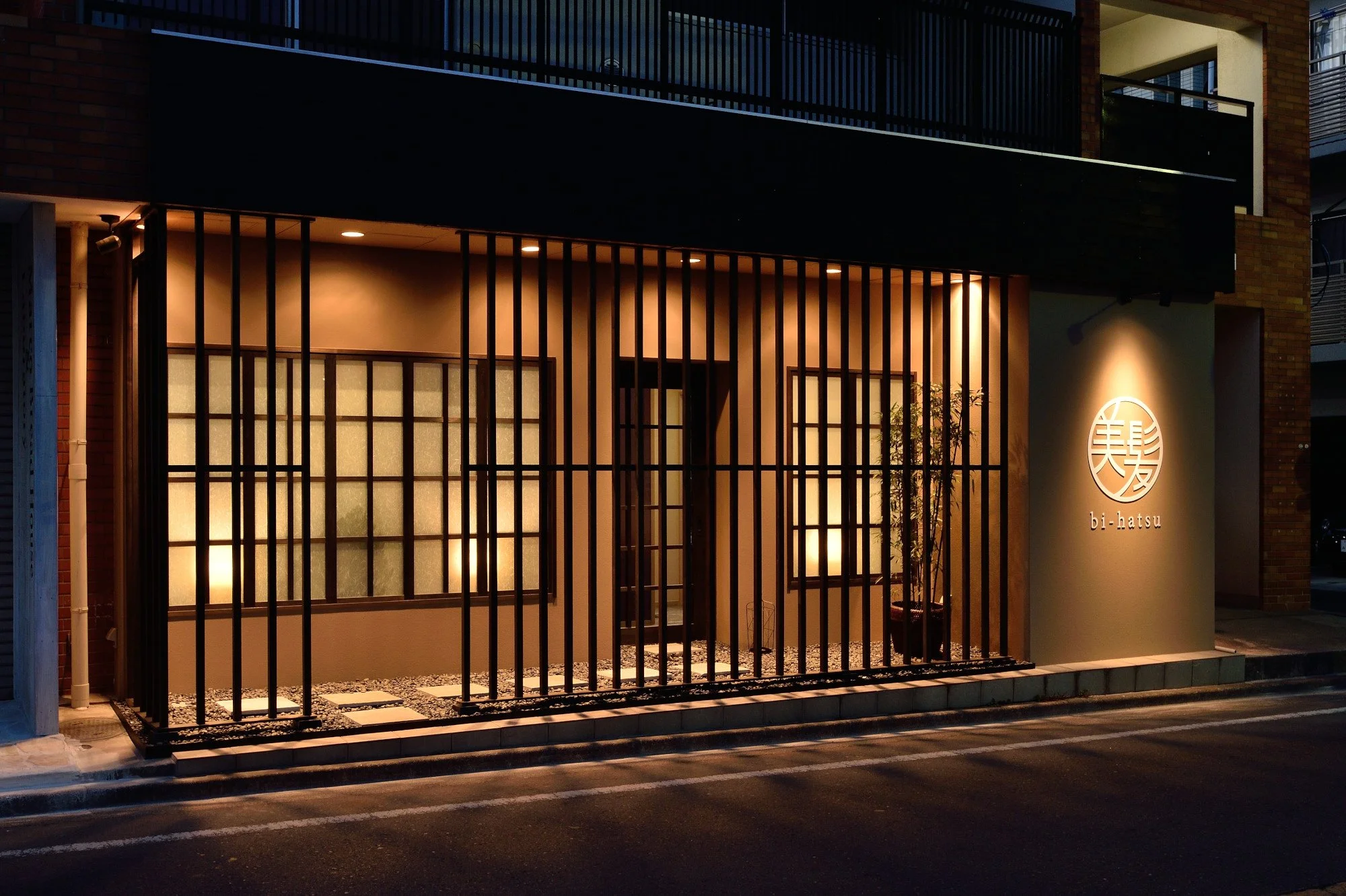 夜の照明に照らされた美容室店舗の外観。木製の格子入りの門と看板には「 bi-hatsu」と書かれている。