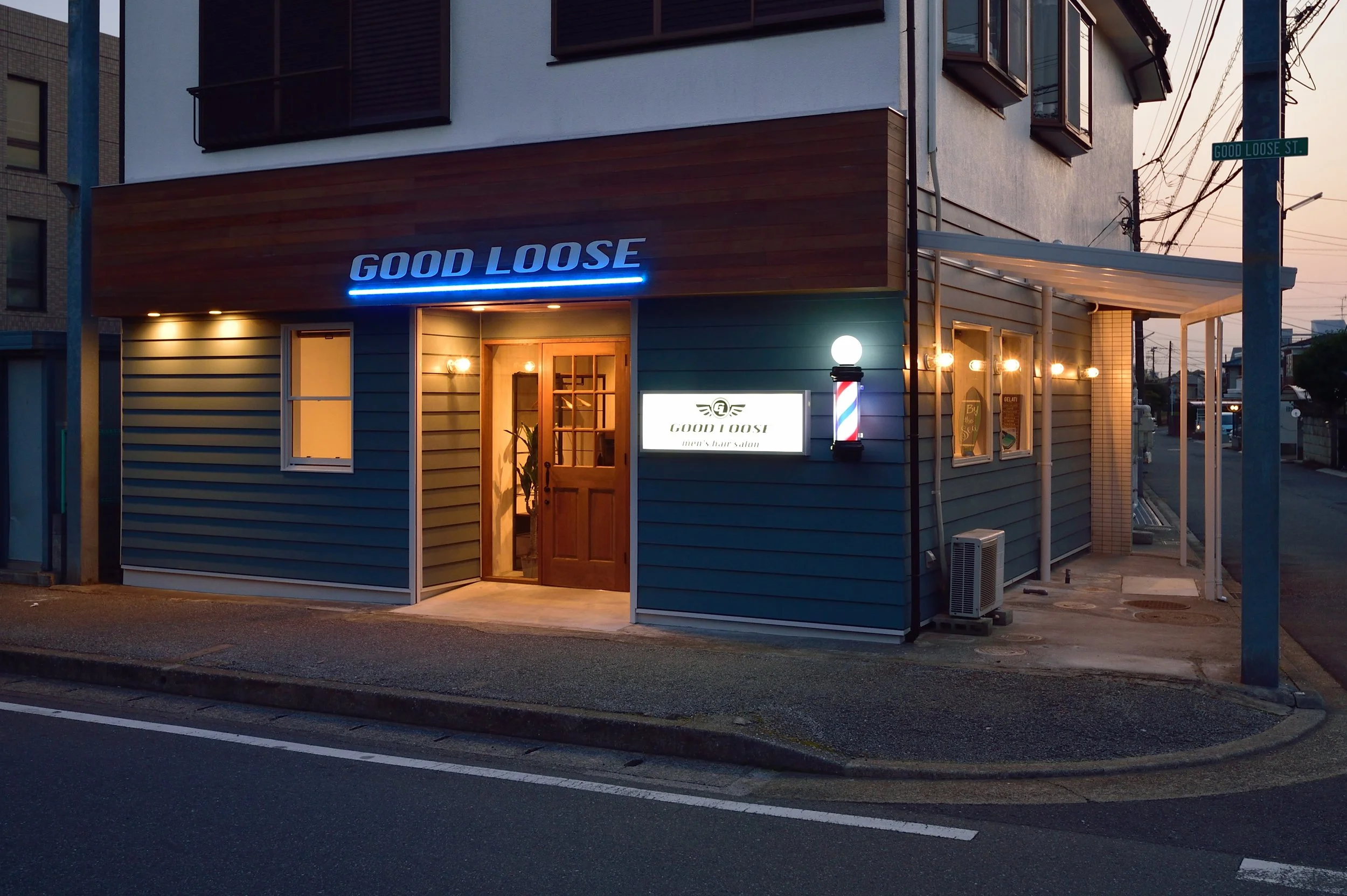 夕暮れ時の青い外壁の美容院『GOOD LOOSE』の看板が灯る店の外観。