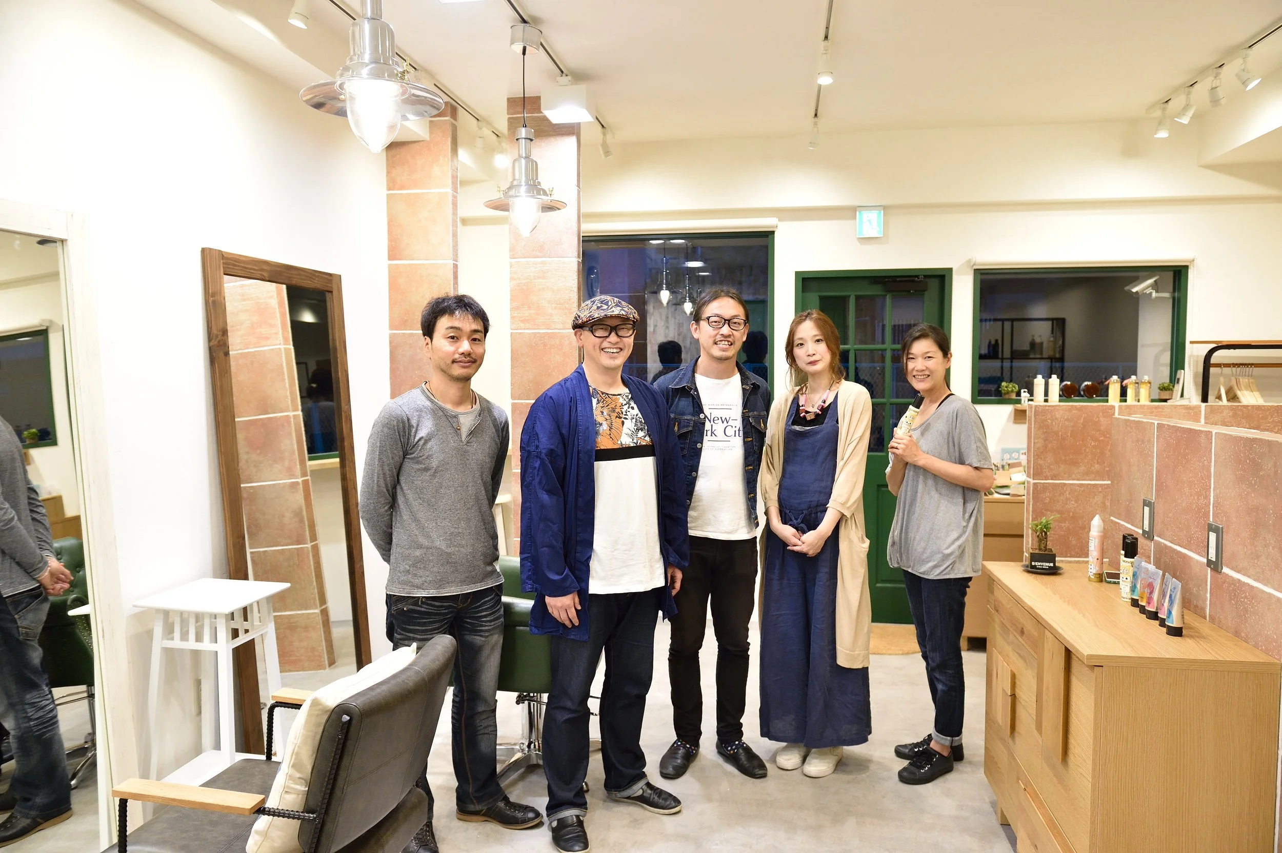 店内で並ぶ男性と女性のグループの写真。背景には緑のドアと出窓、棚に化粧品や小物が置かれている。