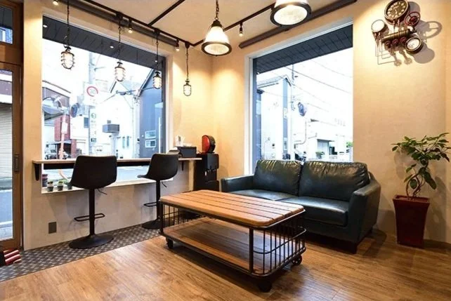 店舗開業インストラクターbh飯島由敬のサポートにより、さいたま市北区日進町に2025年5月開業した理容室balmersの開業時撮影写真。窓側にカウンター席と椅子、中央に木製のテーブルとソファ、壁に時計と観葉植物が飾られている。