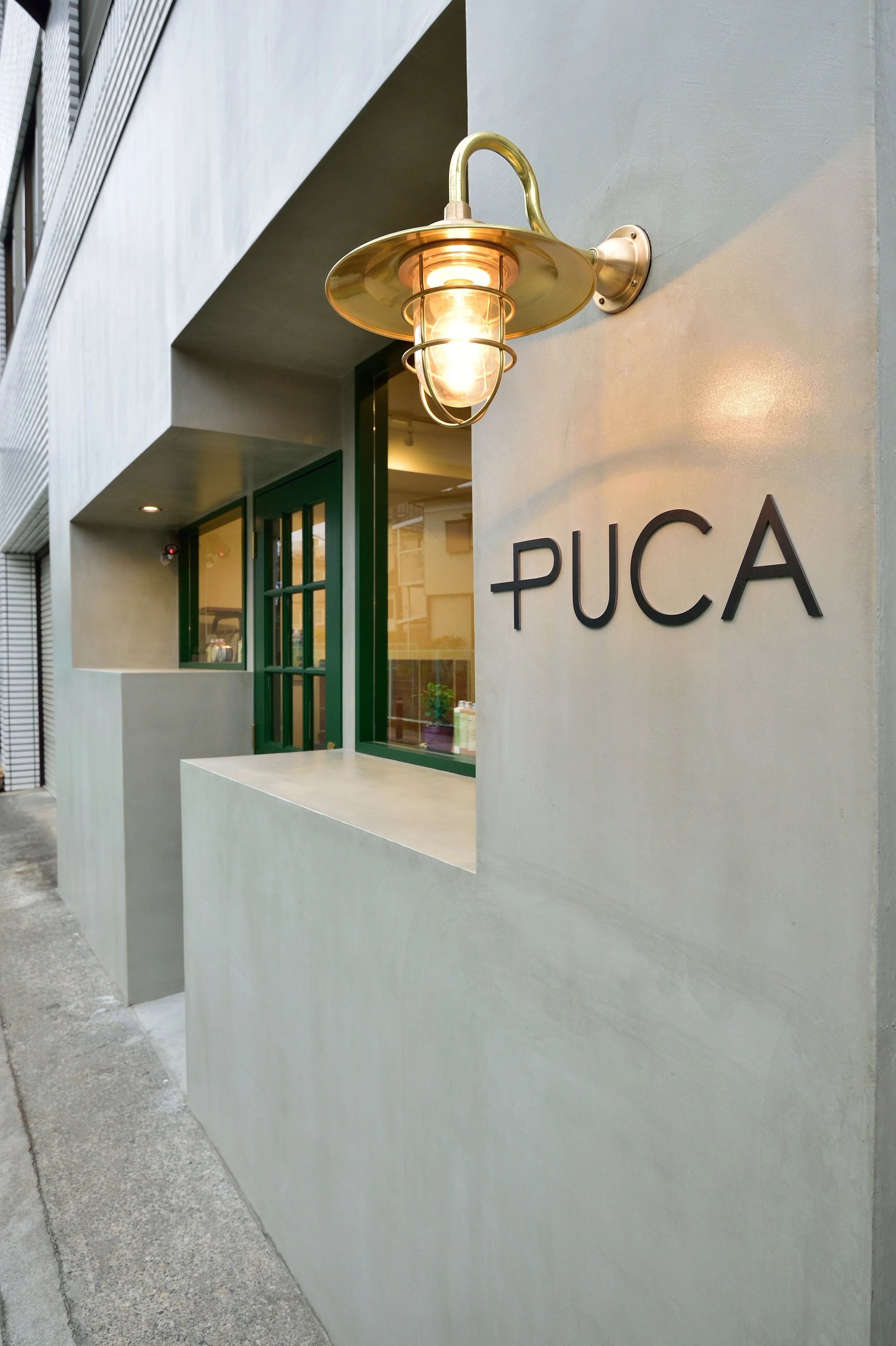 白い壁に取り付けられた金色の外灯と、その下にある黒文字の看板に「PUCA」と書かれている建物の外観。